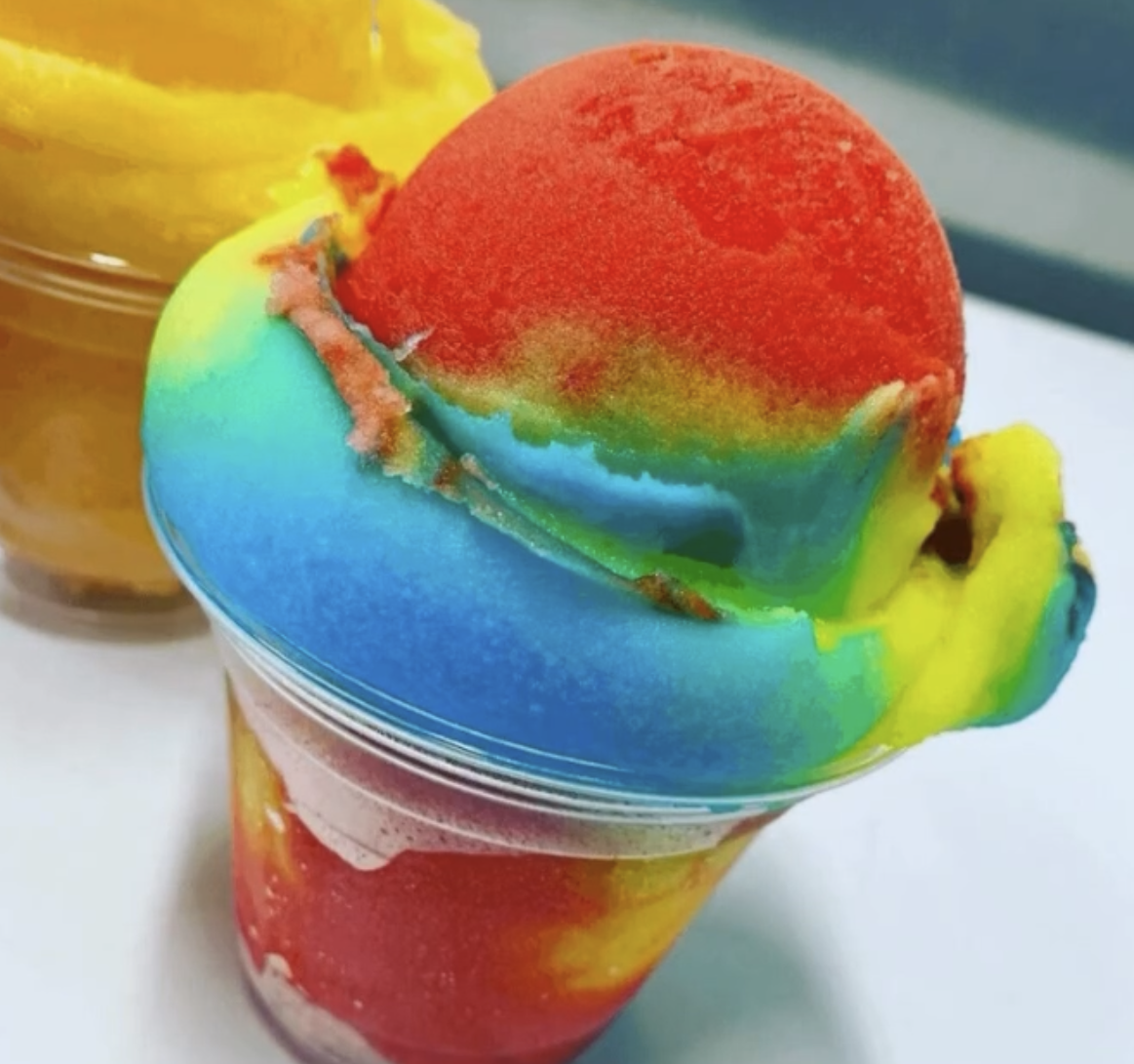 A rainbow-colored shaved ice with a scoop of red ice cream on top, in a clear plastic cup.