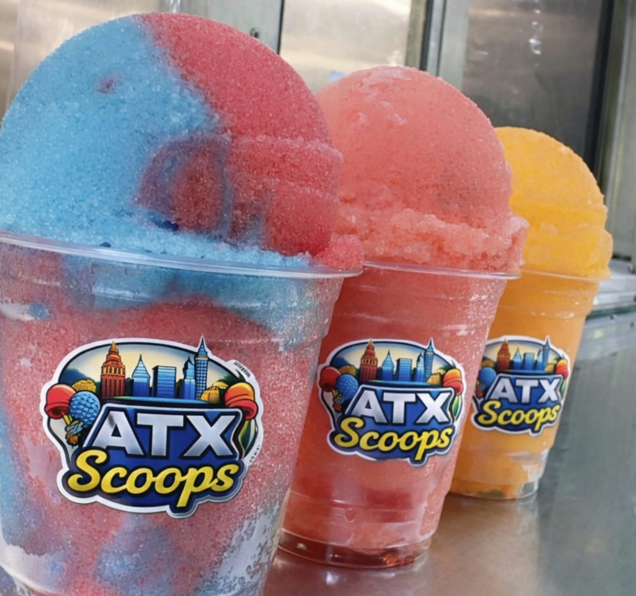 Three cups of colorful snow cones with ATX Scoops stickers, in blue, pink, and yellow flavors, displayed on a counter inside an ice cream shop.