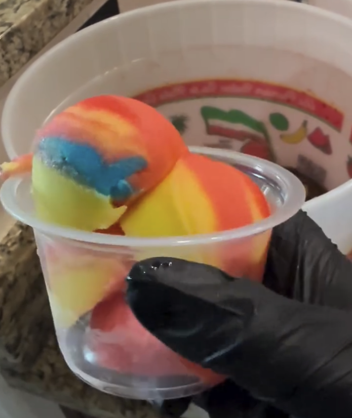 Colorful rainbow ice cream in a small clear plastic cup, with a larger cup of a beverage in the background.