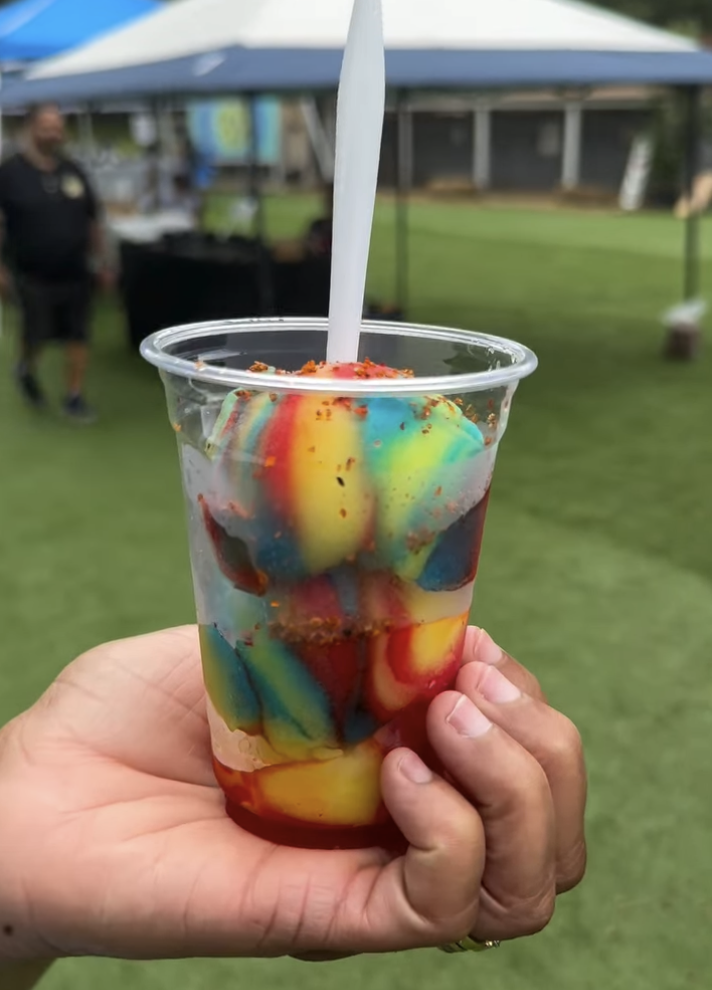 A clear plastic cup filled with colorful, swirled ice cream or popsicle pieces with a straw, held by a person's hand outdoors at a fair or festival with a grassy area and tents in the background.
