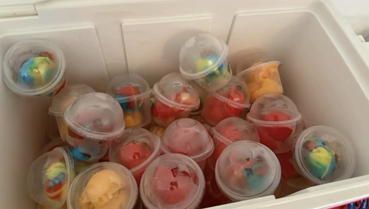 Colorful snow cones with rainbow syrup in clear plastic cups with dome lids inside a white cooler.