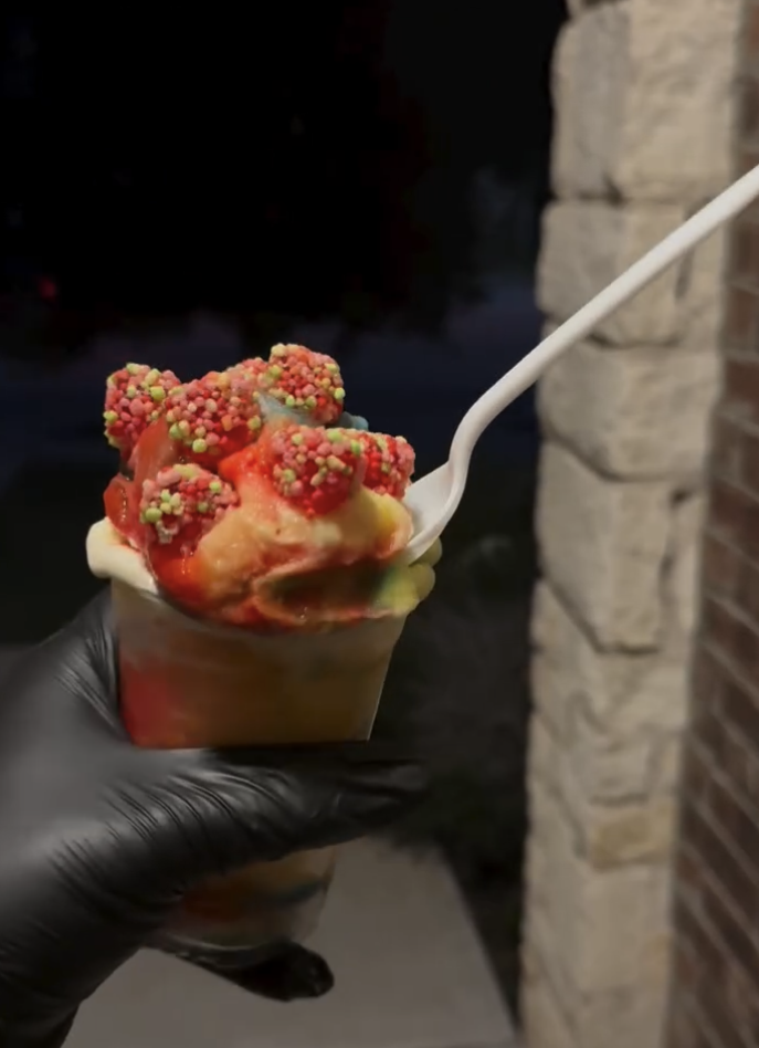 Hand in black glove holding a cup of rainbow sherbet ice cream topped with colorful sprinkles and a white spoon, against a dark and brick background.