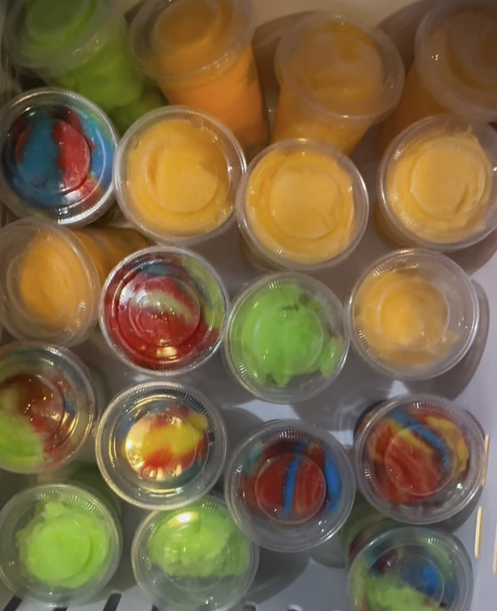 Collection of small, clear plastic cups filled with colorful liquids, some with swirled or marbled appearances.