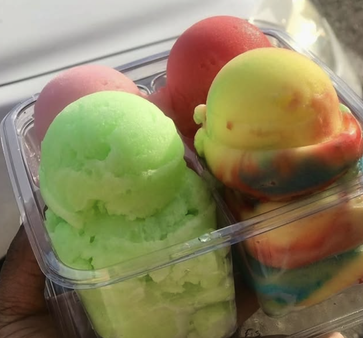 Colorful ice cream scoops in a clear plastic container, featuring green, pink, red, and rainbow swirled flavors.