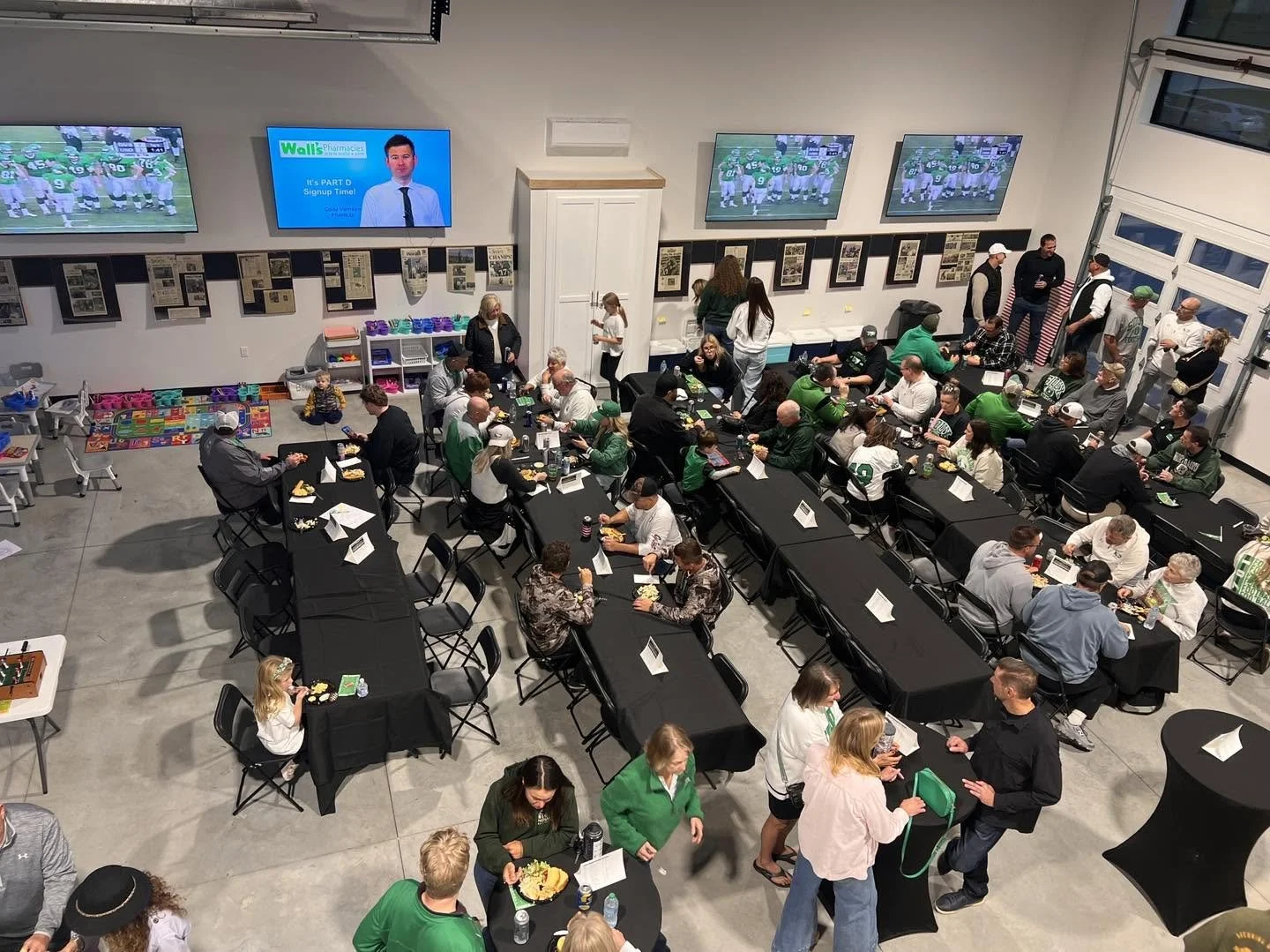 A filled indoor sports watch party with people dining at black-clothed tables, TVs showing a football game, and sports memorabilia on the walls