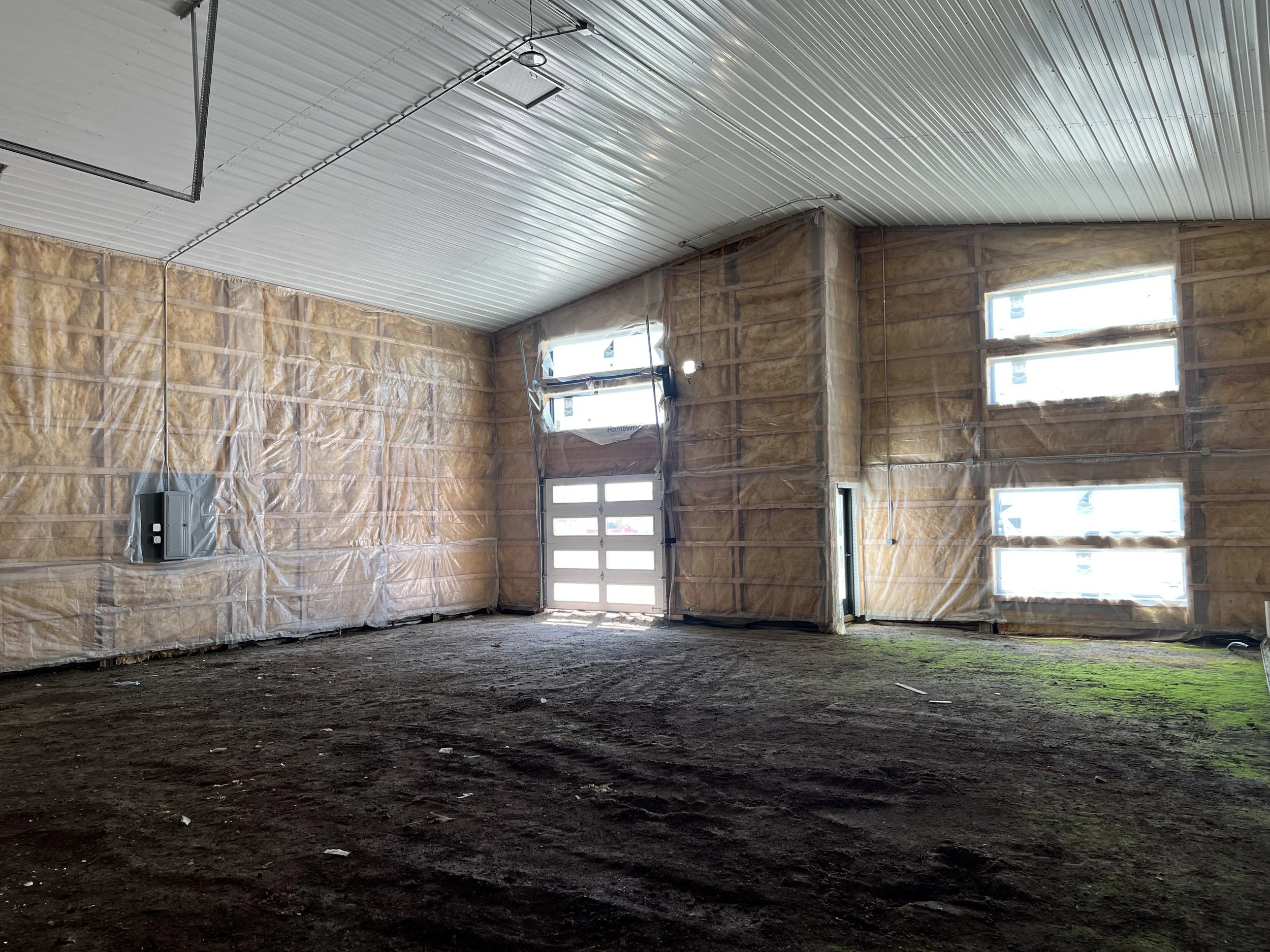 An interior of The Event Bays under construction, with exposed insulation in the walls, a partially finished ceiling with metal panels, and a concrete floor with dirt and some grass growing.