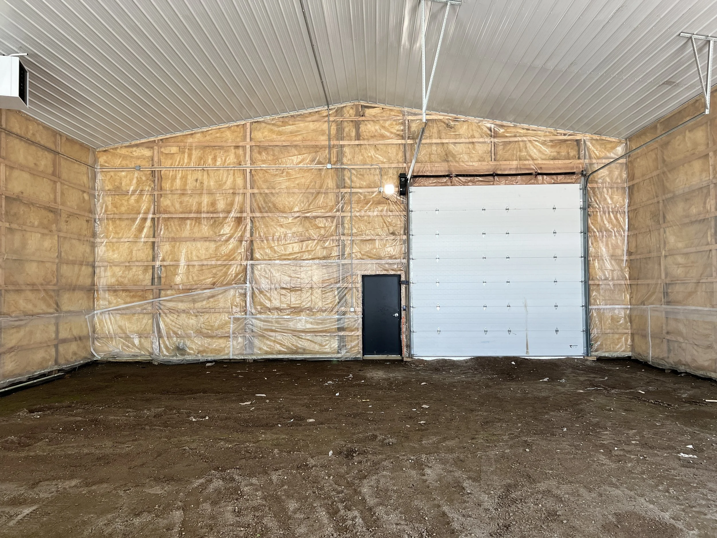 Interior of The Event Bays under construction, with insulation and framing visible, a white garage door, a black door, and a concrete floor.