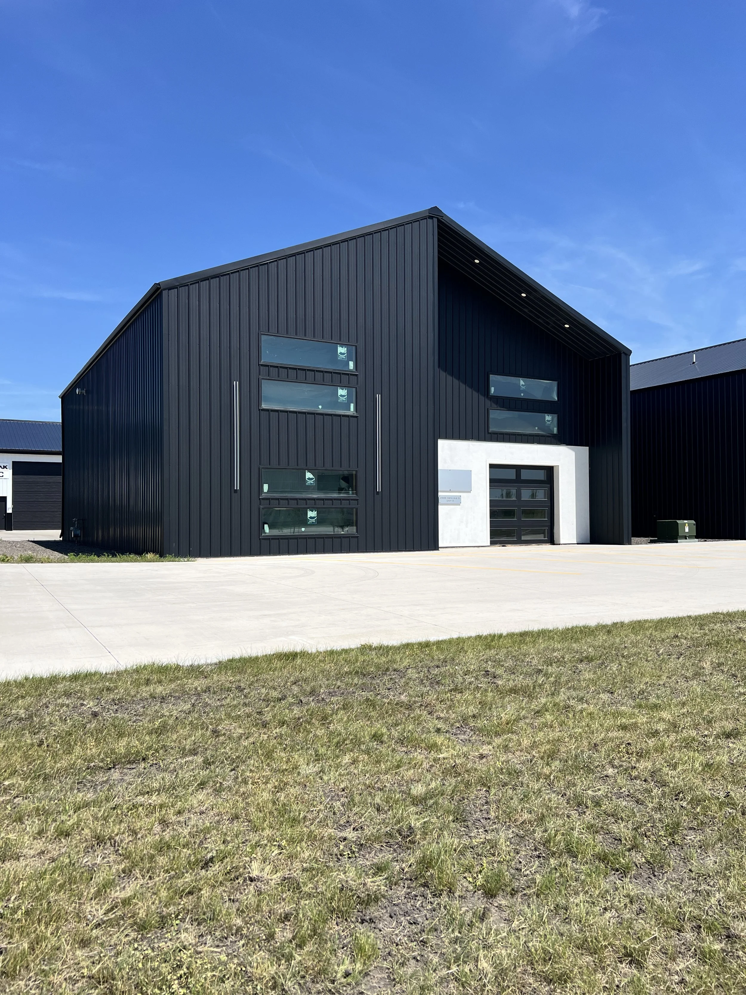 Modern black building with several horizontal windows, set against a blue sky with sparse clouds and a patch of grass in the foreground.