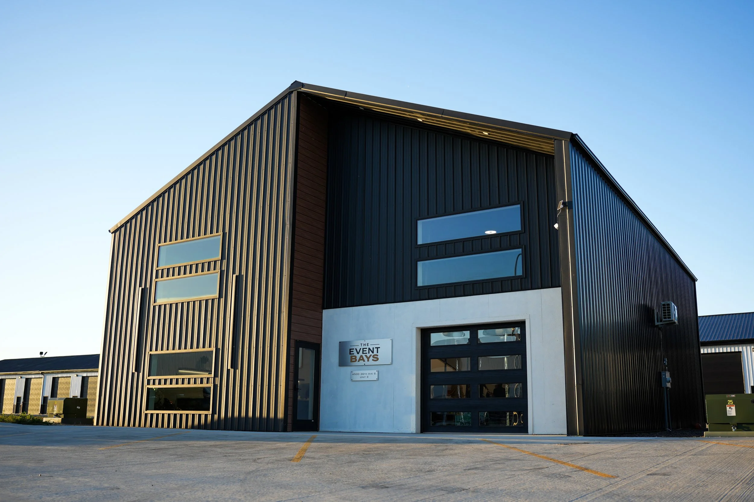 Exterior of The Event Bays event venue, a modern black corrugated-metal building with large windows and garage doors under a clear blue sky.