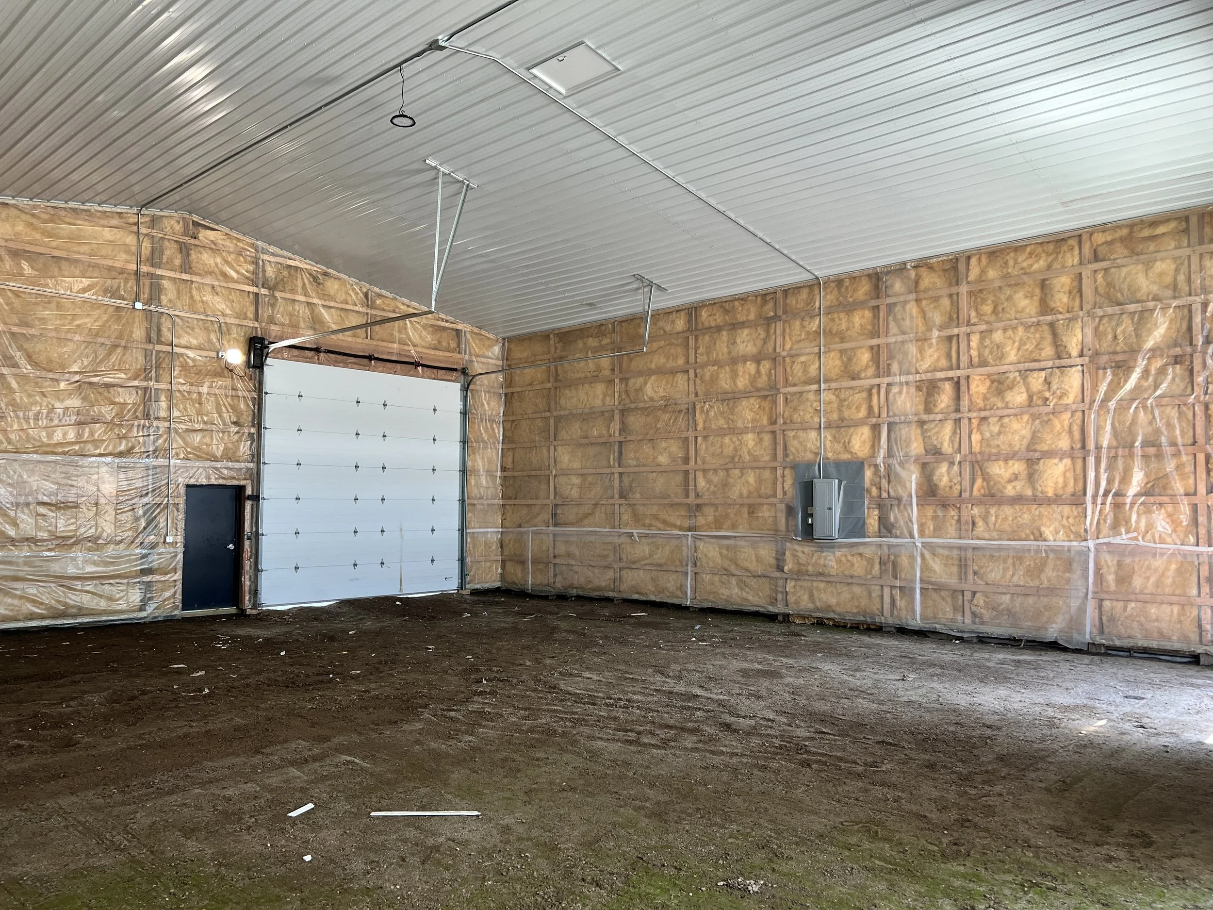Interior of the unfinished Event Bays with insulation on the walls, a white garage door, and a dirt floor.