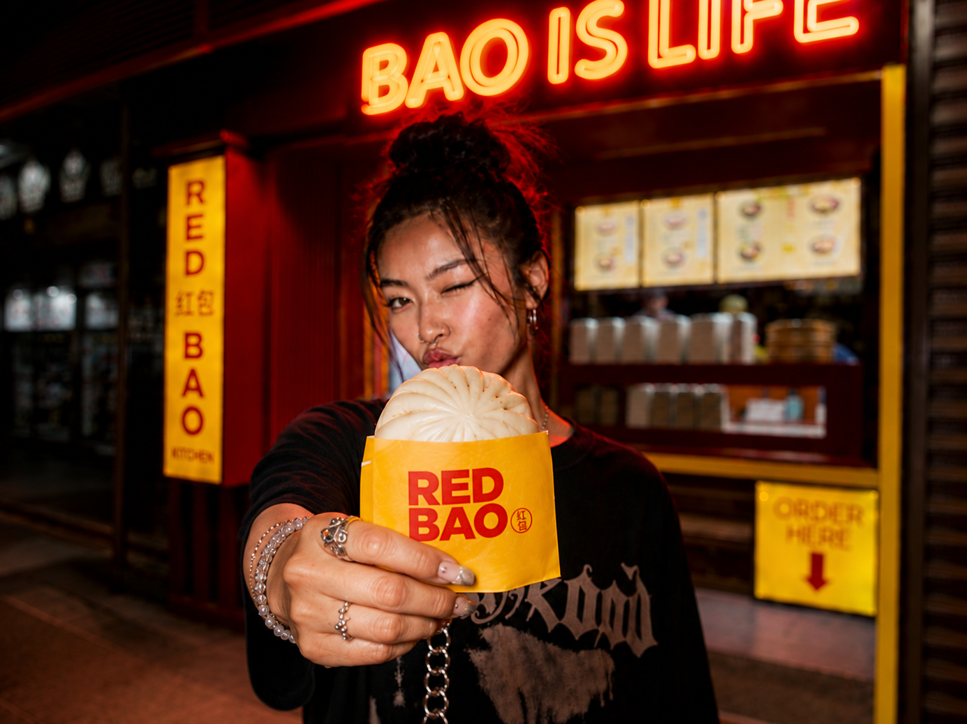A woman with dark hair in a messy bun, wearing rings and earrings, holding a steamed bun in front of her at a food stand. The stand has bright yellow and red signage with the words 'RED BAO' and 'BAO IS LIFE' at night.