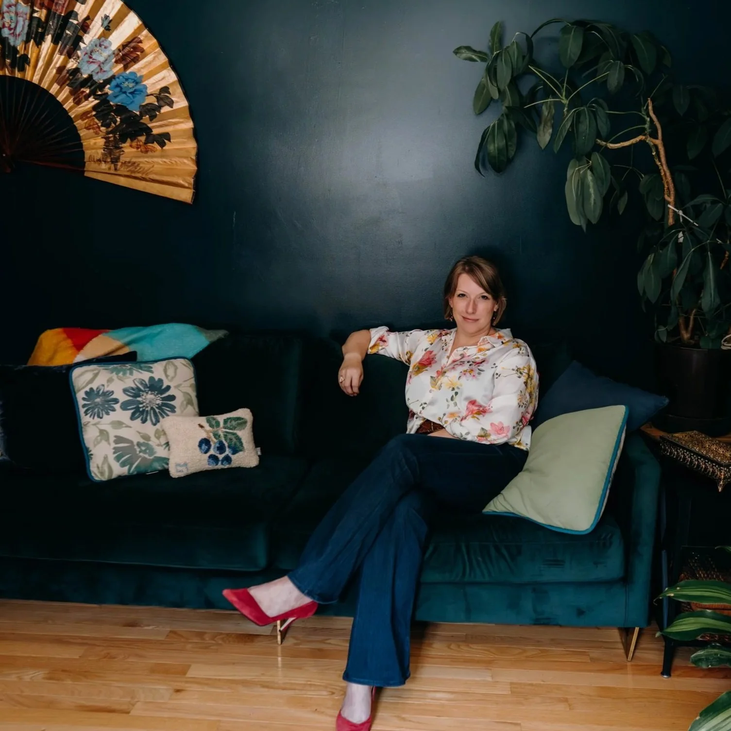 Beth sitting on a teal velvet sofa in her office surrounded by colorful decor and plants.