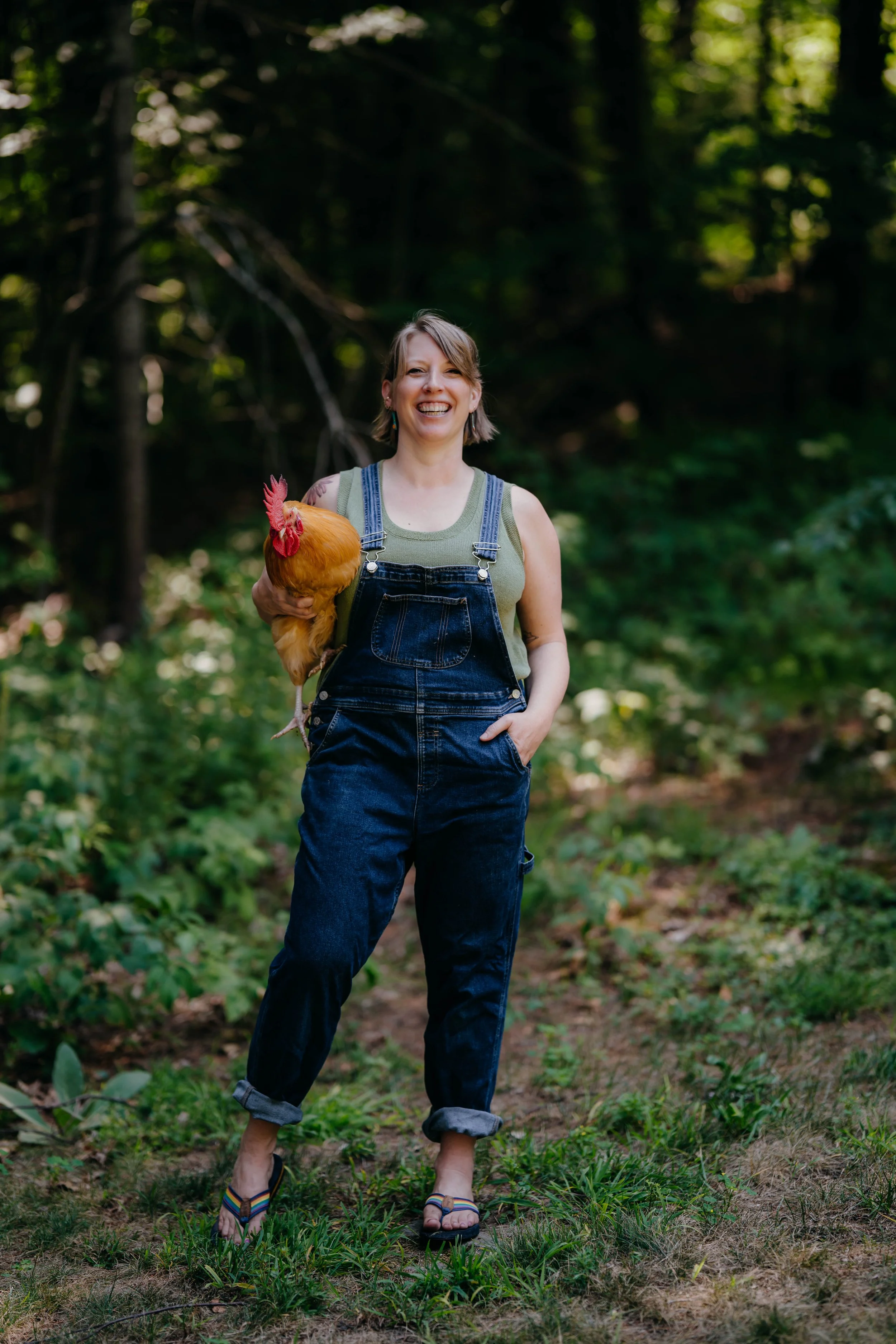 Beth standing in a wooded area holding a chicken, smiling and wearing casual clothing including overalls and flip-flops.