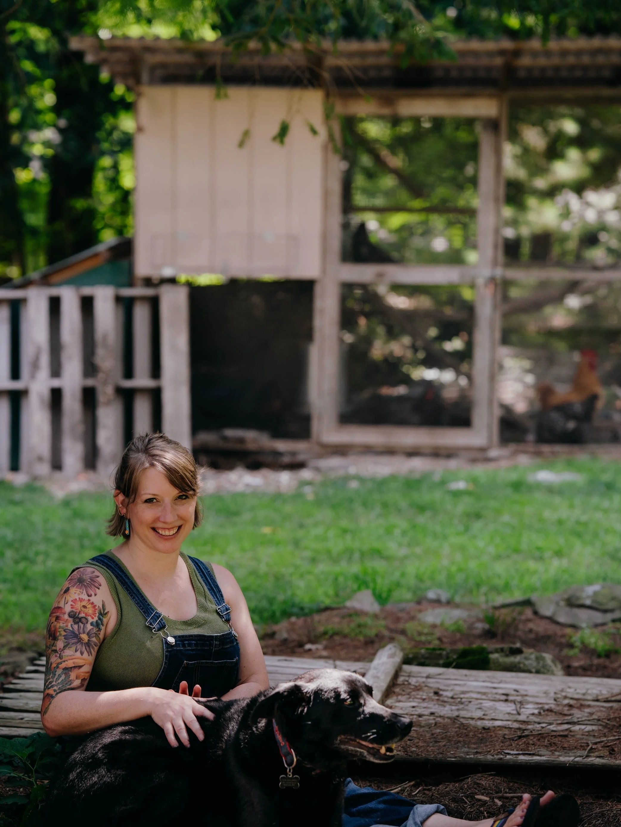Beth wearing denim overalls sitting outside with her dog Cooper with the chicken coop in the background.