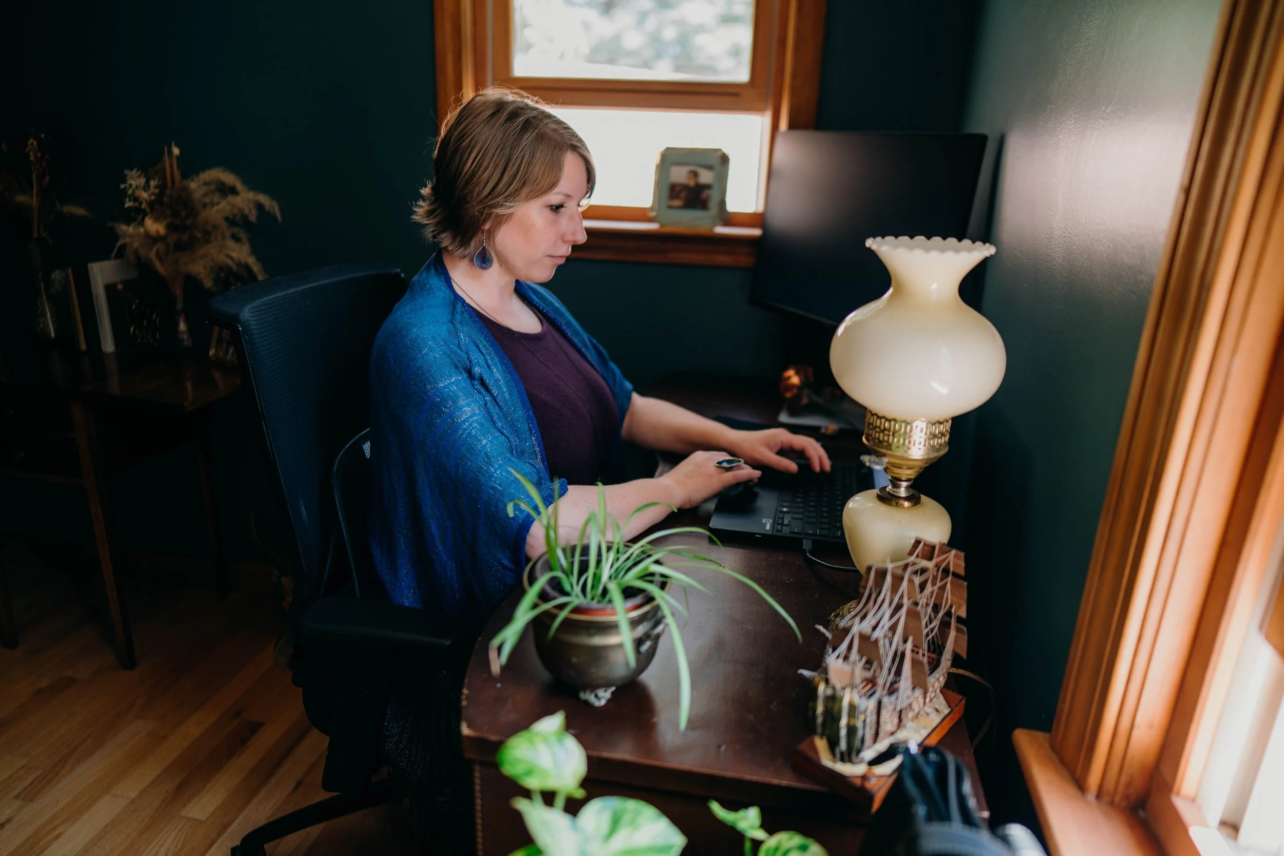 Beth sitting at her desk working on a laptop surrounded by plants and colorful decor.
