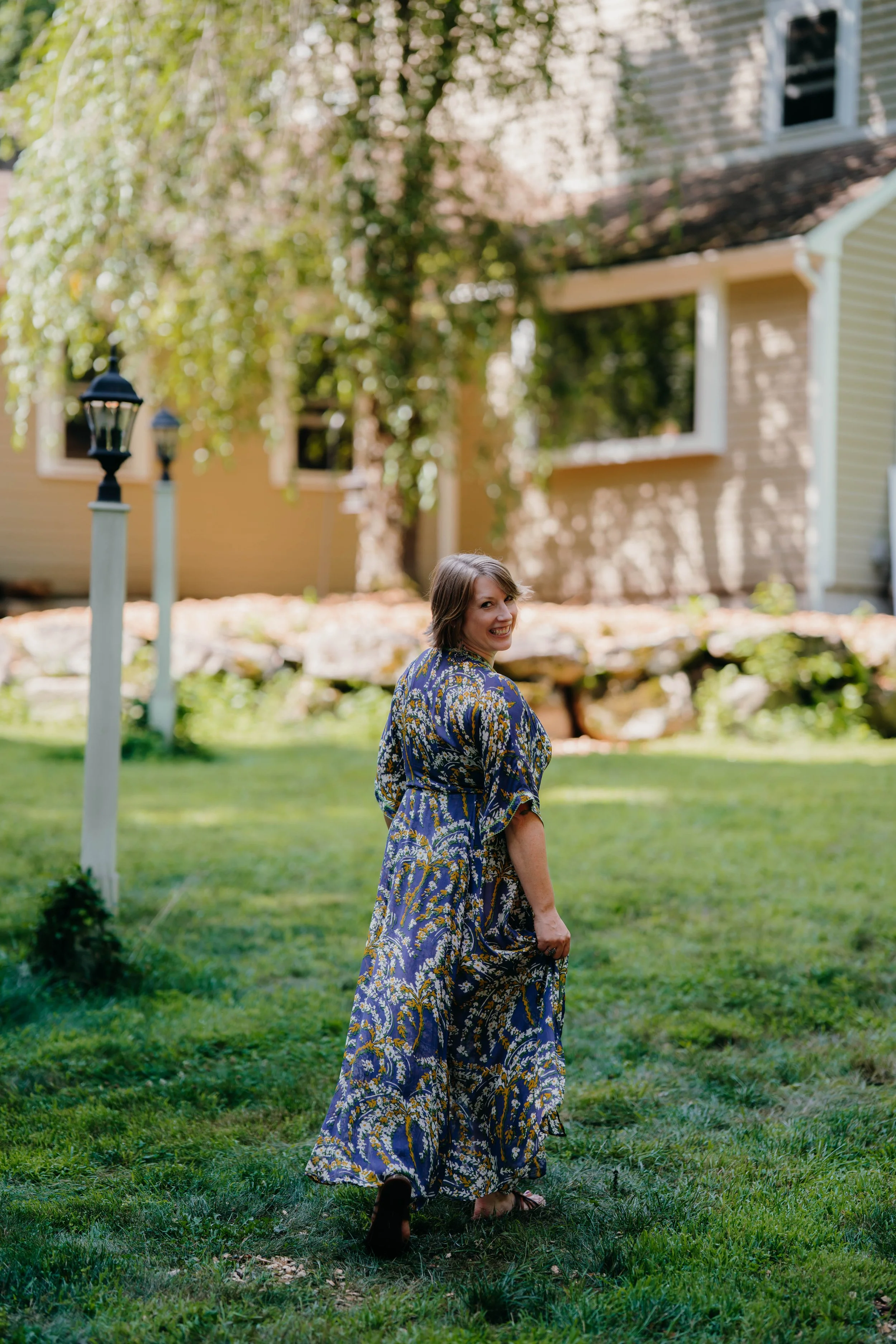 Beth in a patterned blue, yellow, and white dress walking on grass in her backyard with trees, a house, and lamp posts in the background.