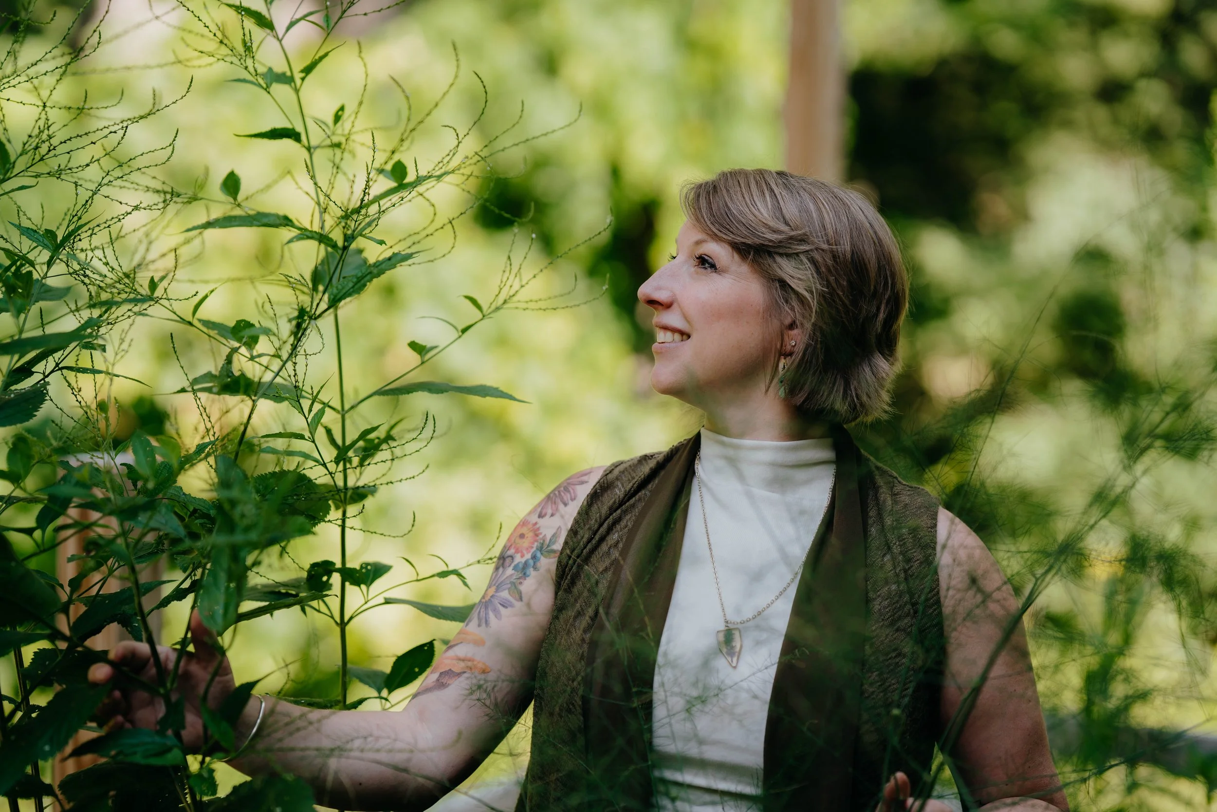 Beth smiling and looking up, standing amidst green foliage in her garden.