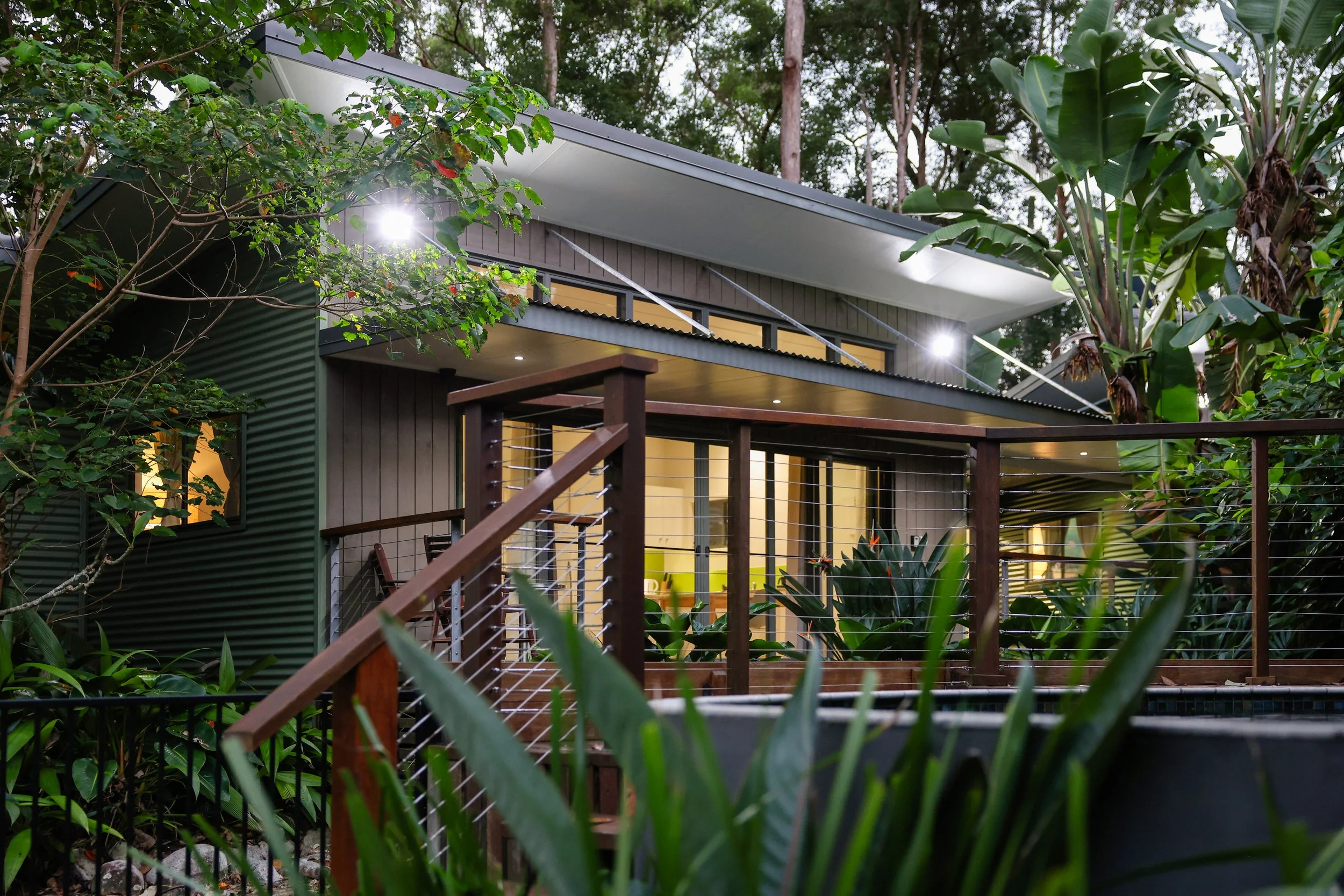 Modern house surrounded by lush green trees and plants, with a balcony and outdoor stairs, at dusk with interior lights on.