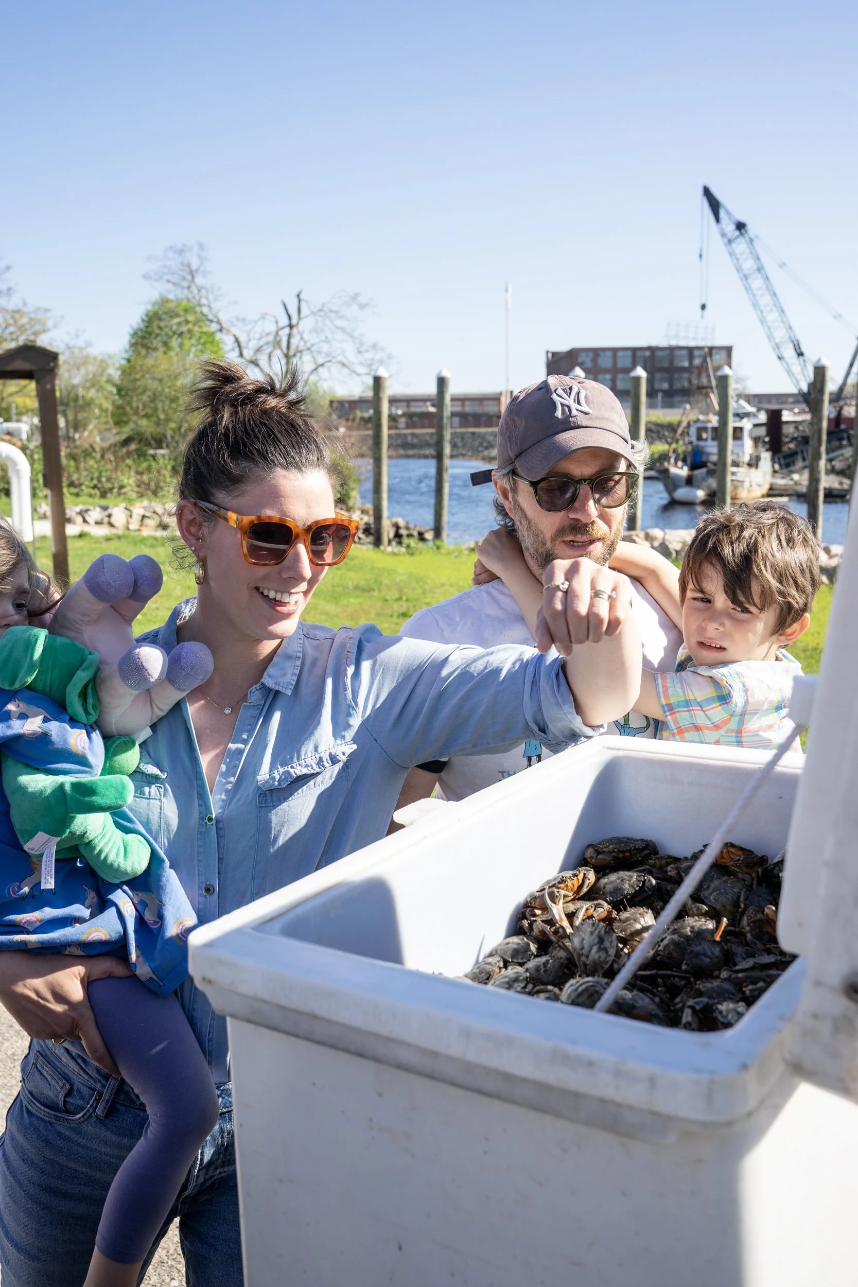 Family at a dockside seafood farm, holding a large container of freshly harvested oysters or shellfish, with boats and a crane visible in the background.