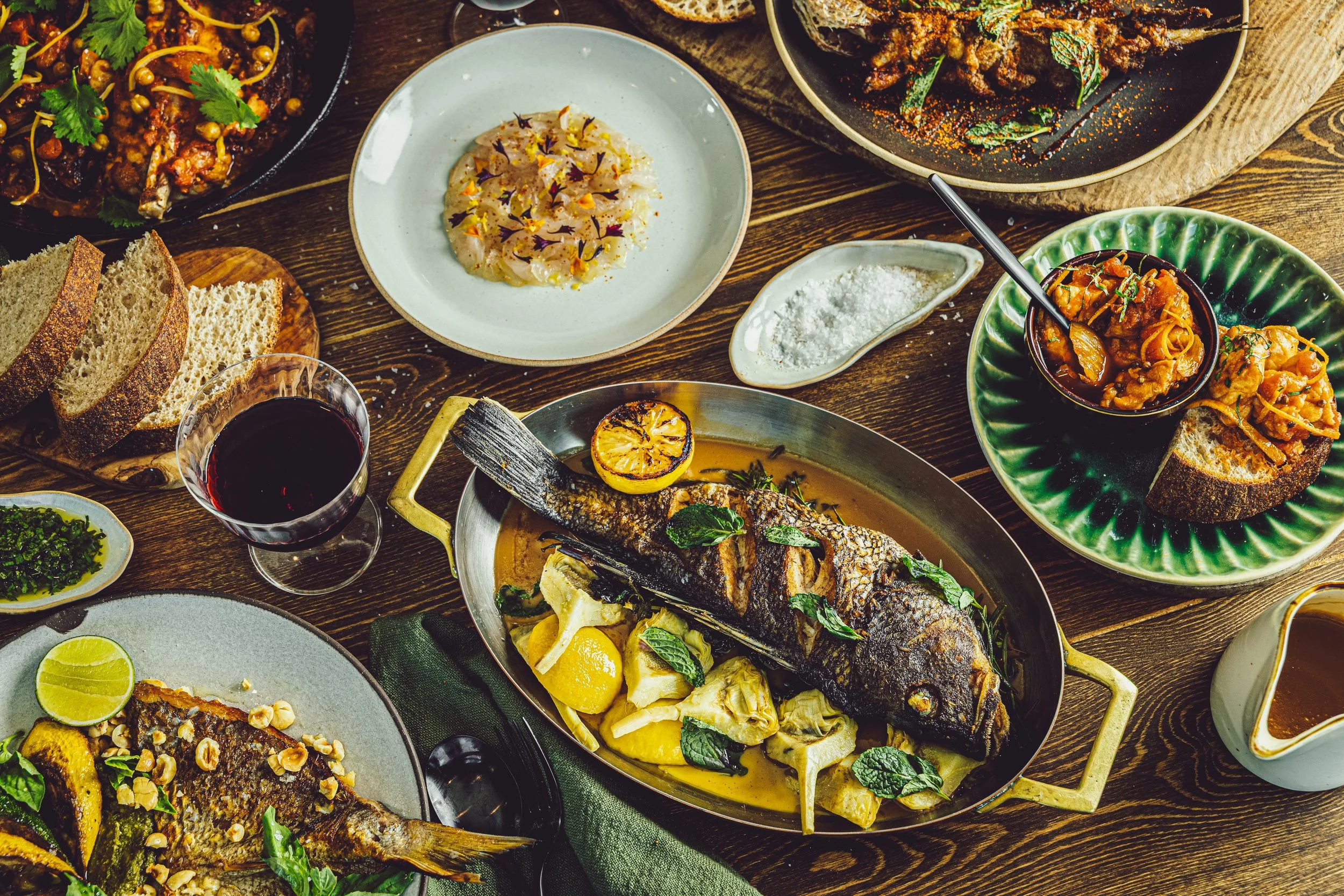 Assorted dishes including grilled fish with lemon, vegetable-based salads, bread slices, red wine, and various spices and garnishes on a wooden table.