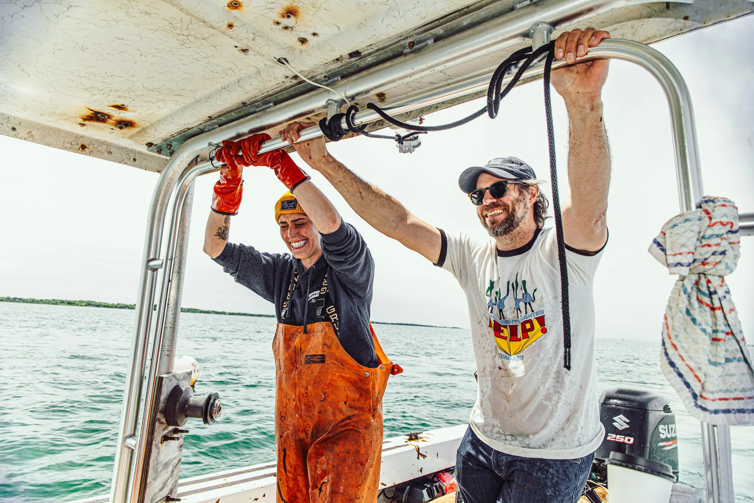 Two people on a boat holding onto a metal rail, smiling and wearing casual clothes. One person is wearing a yellow beanie and orange waterproof pants, while the other person is wearing sunglasses, a cap, and a white t-shirt. The boat is on water, with a cloudy sky in the background.