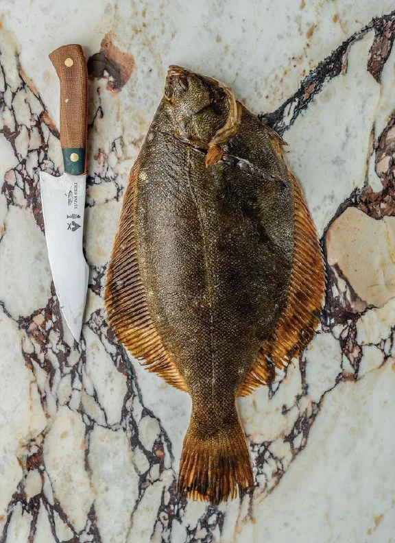 A whole fluke butchery technique, with a chef's knife placed next to it on a marble surface.