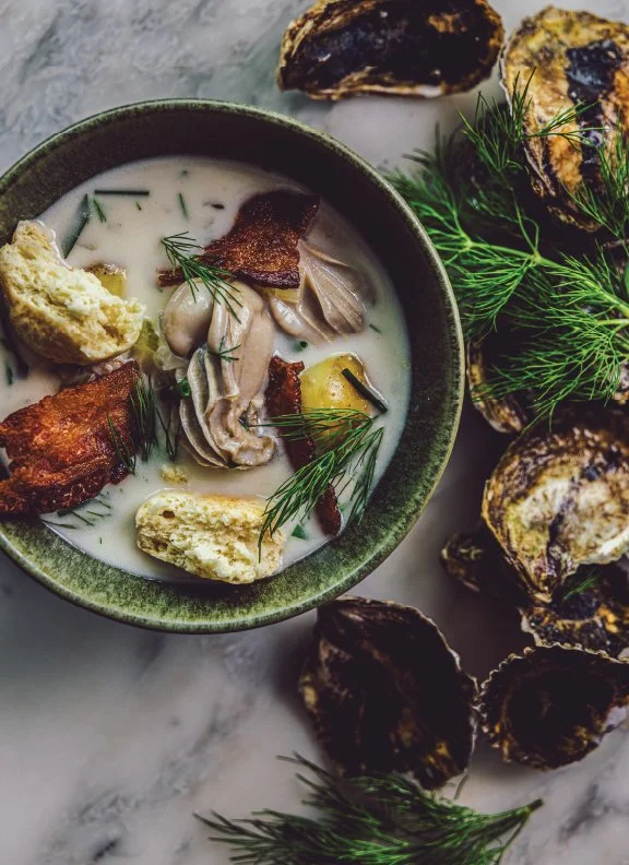 A bowl of New England Oyster Chowder garnished with herbs, served with roasted oysters and dill on a marble surface.