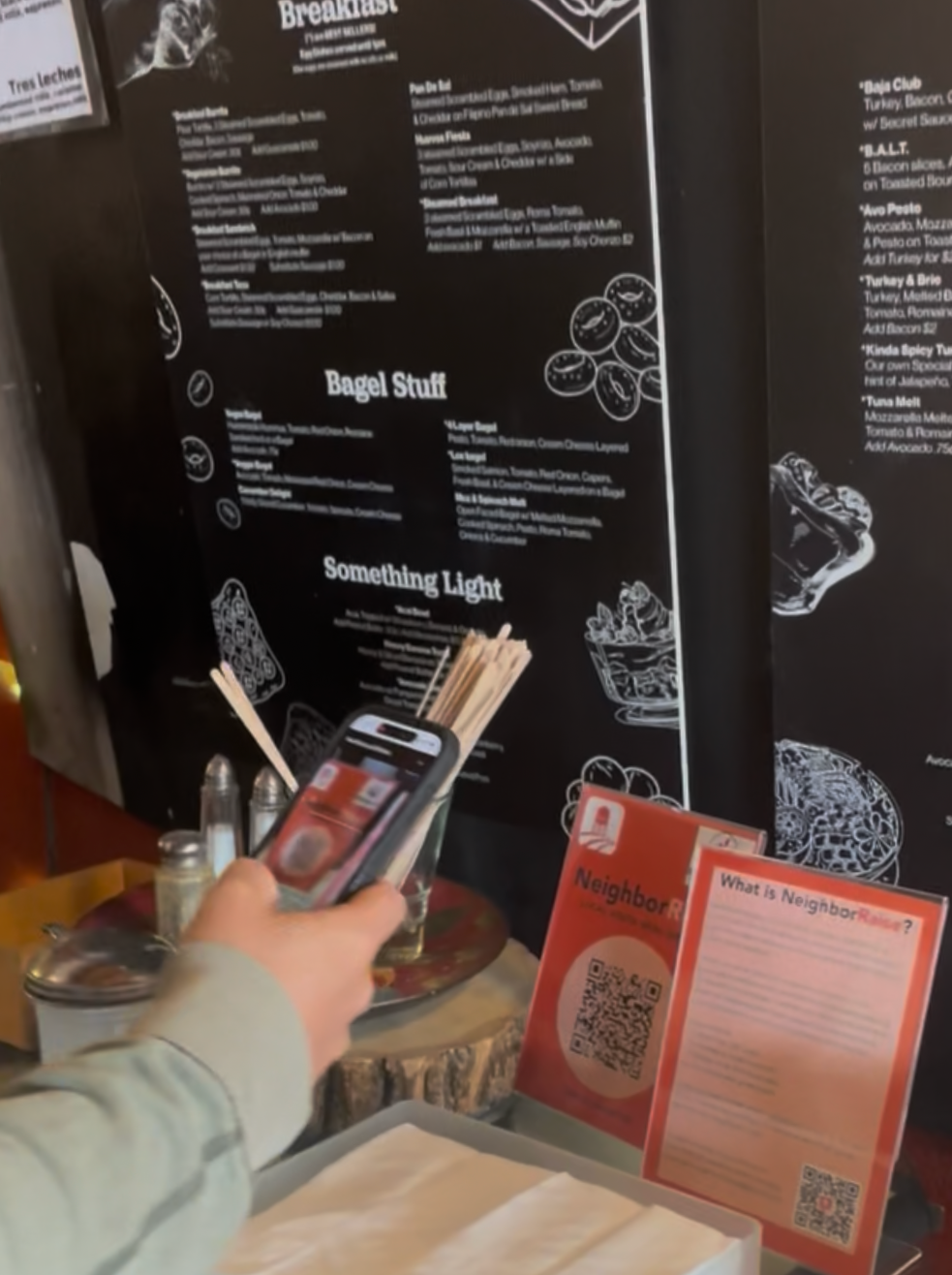 Person holding smartphone in front of a restaurant menu with various food categories and illustrations, including a glass of beer and a bowl of food, on a counter with condiment bottles and napkins.