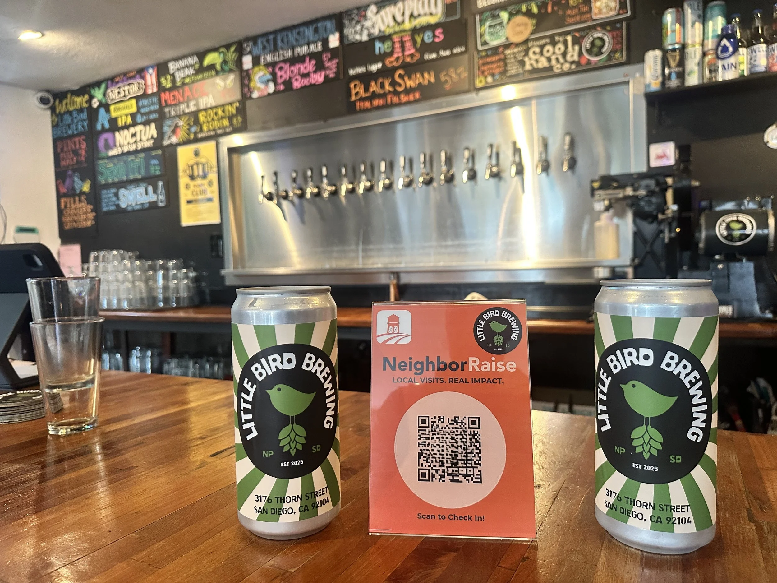 The image shows the inside of a taproom or brewery with a row of beer taps mounted on a stainless steel panel. There are colorful chalkboard-style signs above the taps advertising various beer options. In the foreground, there are two cans of Little Bird Brewing beer and a red check-in QR code card on a wooden table, with a glass and some empty glasses nearby.