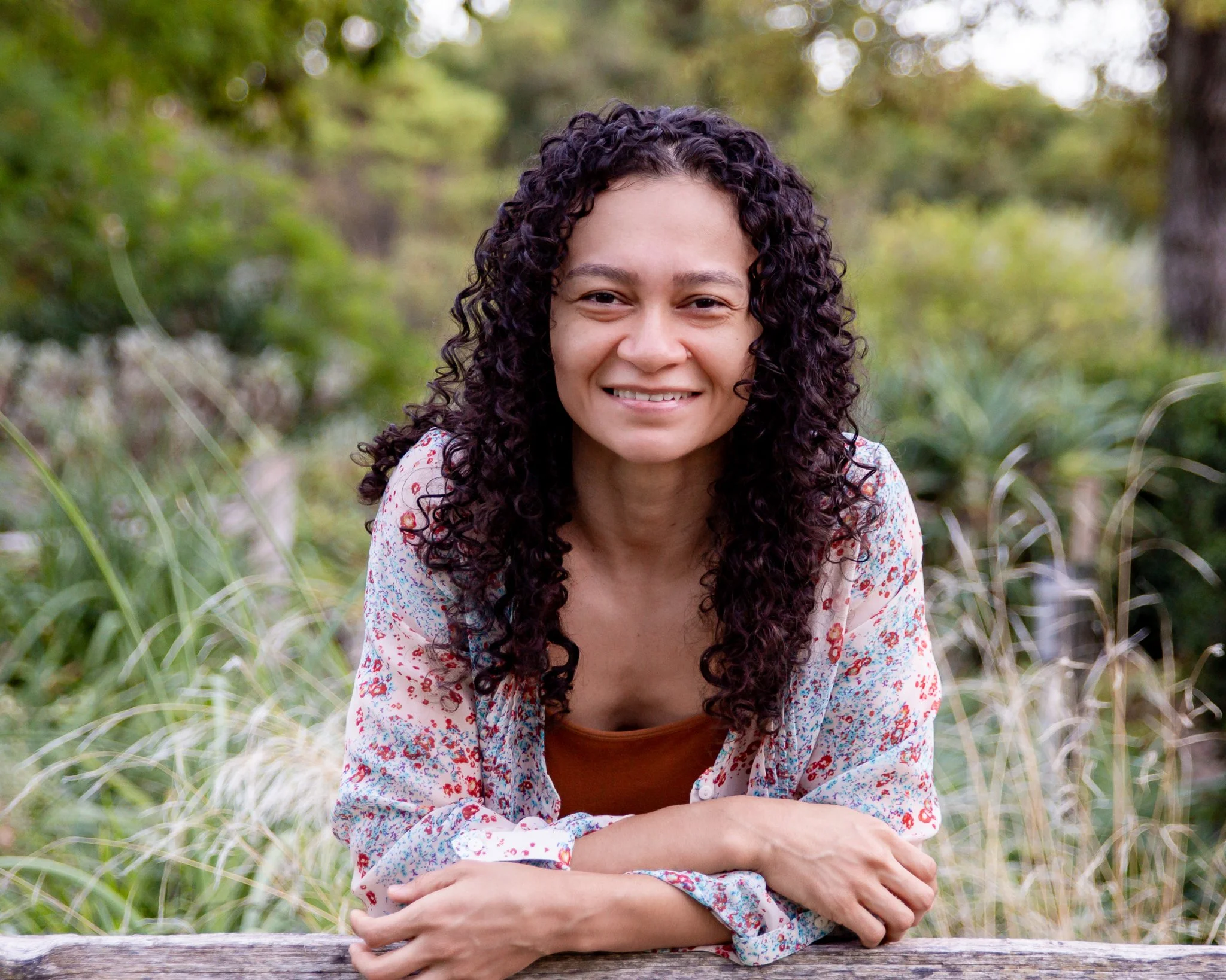 Woman smiling at camera leaning on fence with plants in background. She is a trauma therapist Sacramento