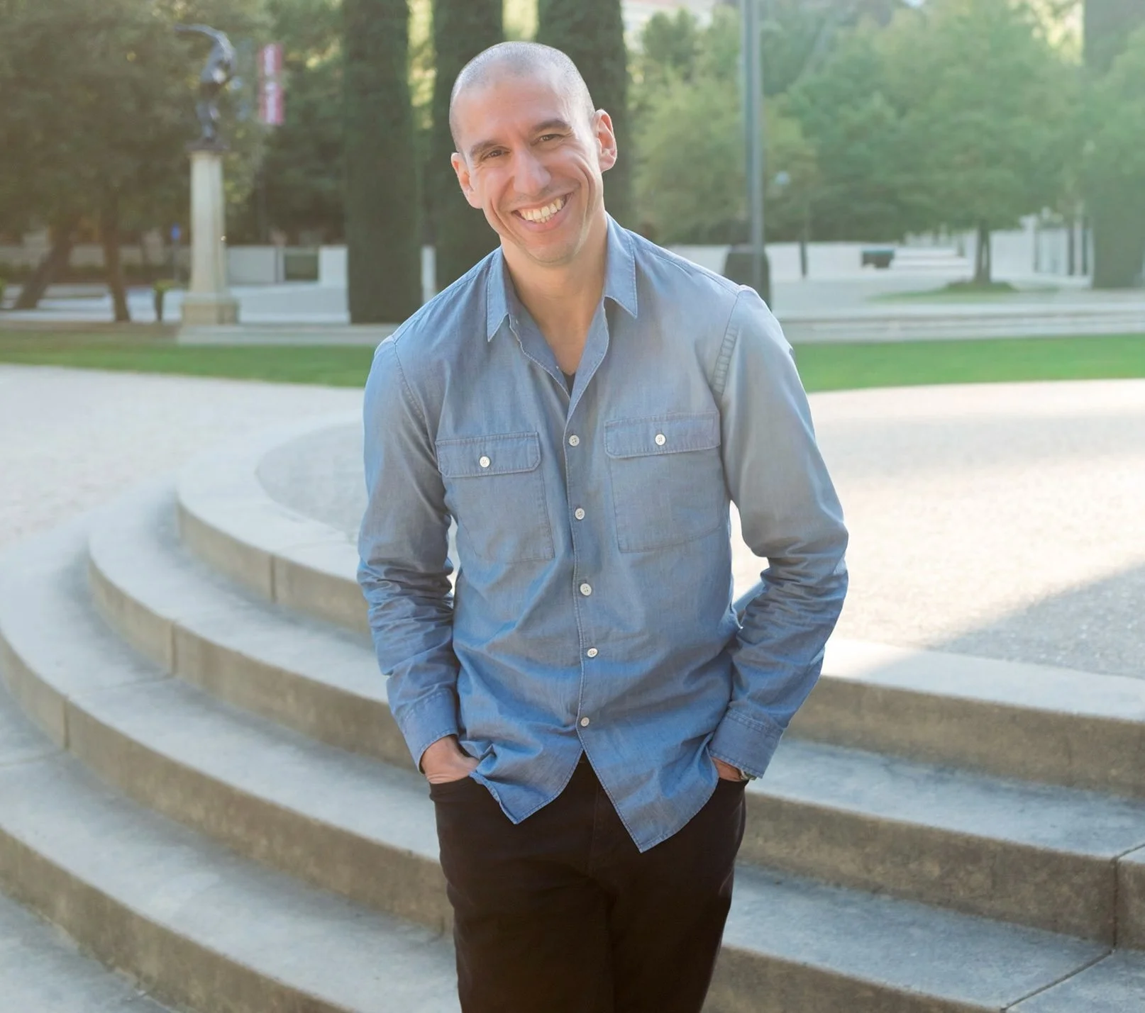 A man wearing a light blue denim shirt and dark pants standing outdoors on stone steps with a park in the background, smiling and looking at the camera.