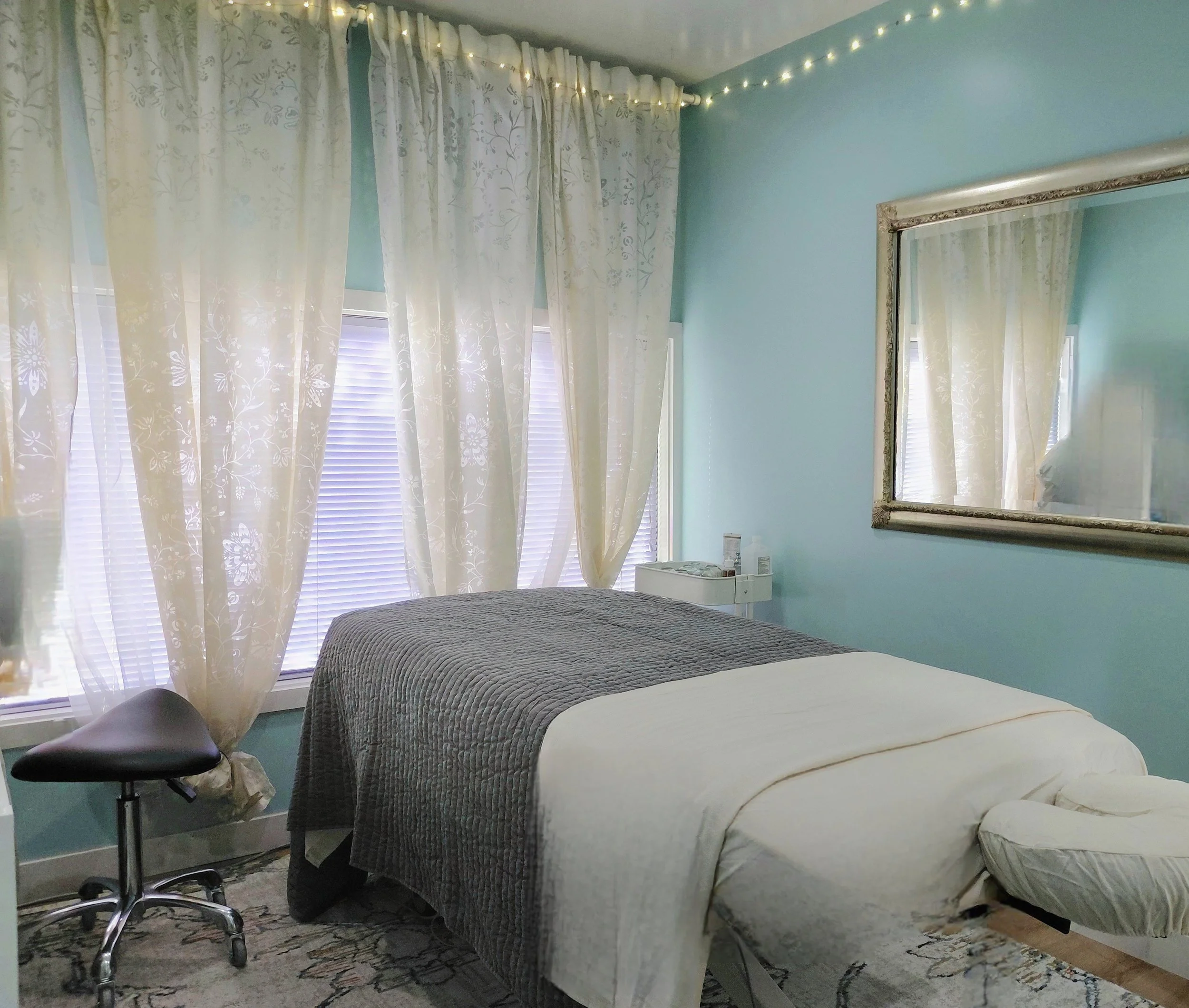 A bedroom with light blue walls, a bed with a dark gray quilt, and a cream blanket. There is a mirror with an ornate frame on the wall. Cream curtains with a floral pattern cover the window, and fairy lights are strung along the ceiling.