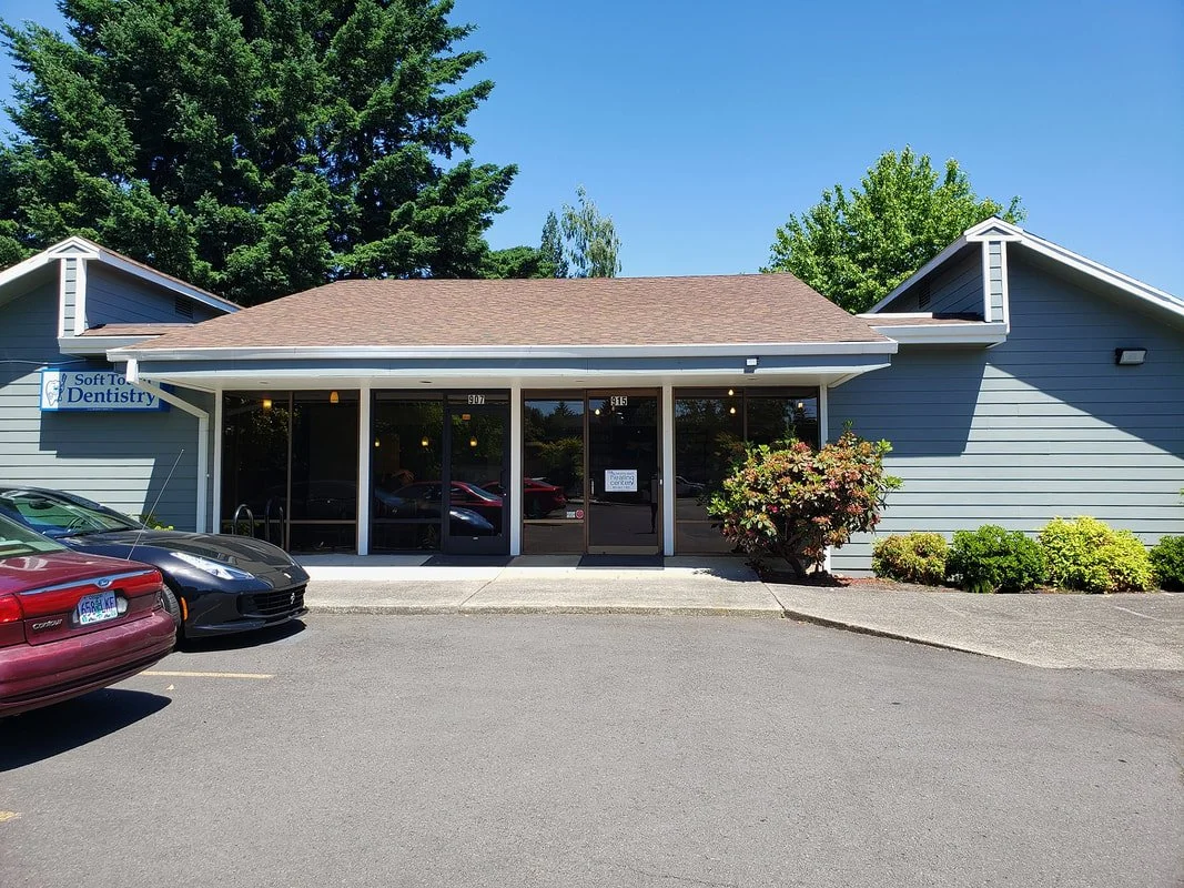 Exterior of a dental clinic with a sign reading 'Soft To' Dentistry, parking lot in front, multiple cars parked, shrubs and trees surrounding the building.