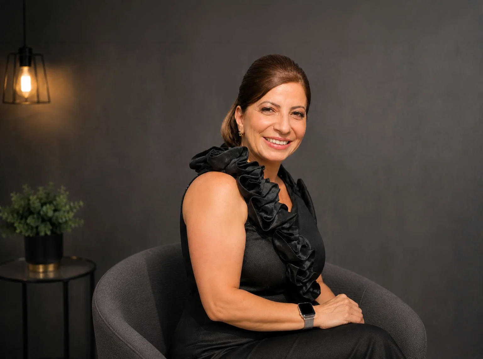 A woman with brown hair styled back, wearing a black dress with ruffled details on the shoulder, sitting on a gray chair in a dimly lit room with dark gray walls, a black side table with a potted plant, and a hanging light fixture in the background.