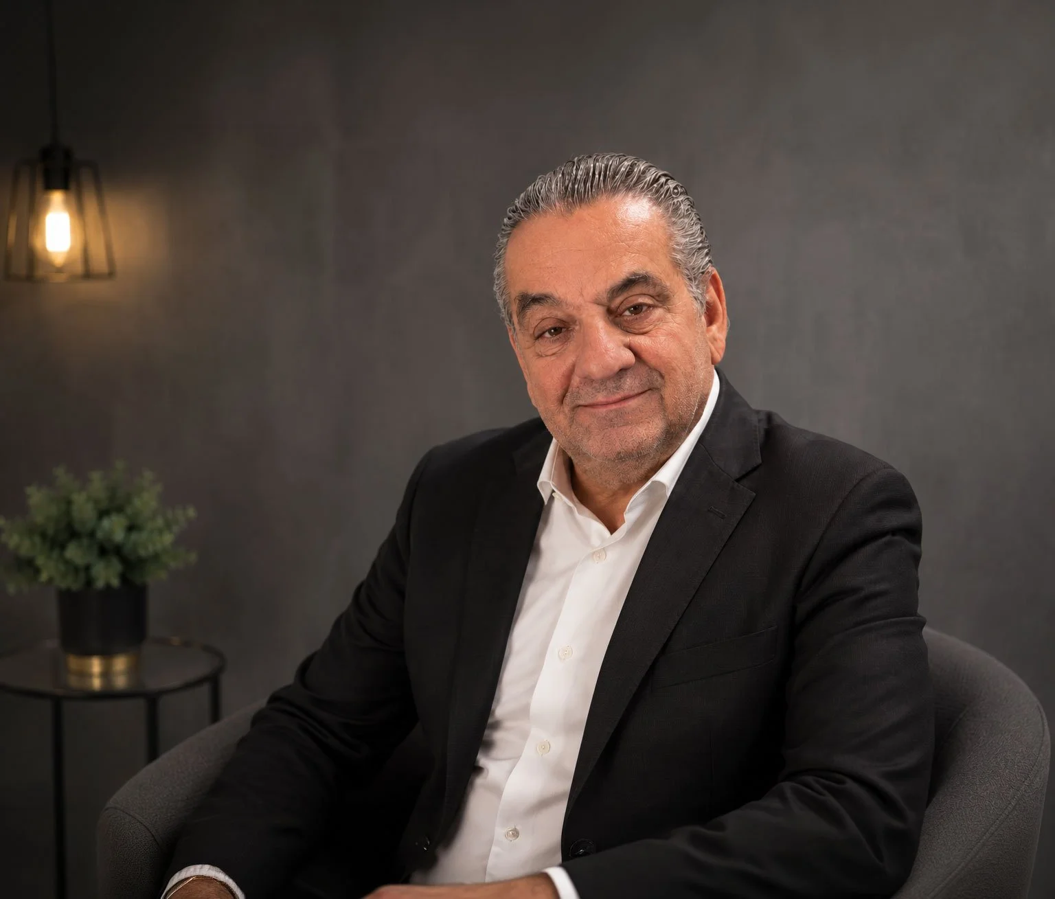 A middle-aged man with gray hair, wearing a black suit and white shirt, sitting on a gray chair in a dimly lit room with a dark textured wall, a small side table with a potted plant, and a warm light fixture in the background.