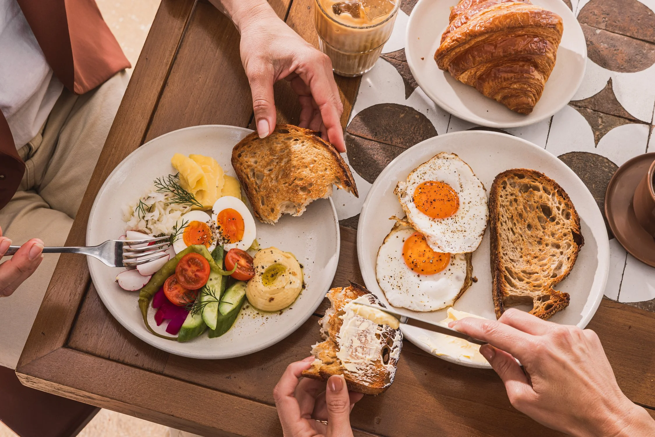 A breakfast scene with multiple plates of food on a wooden table. There are fried eggs, sliced tomatoes, cucumbers, pickles, mashed potatoes, and radishes, along with toasted bread and a croissant. Several hands are reaching for the food, some holding bread slices.