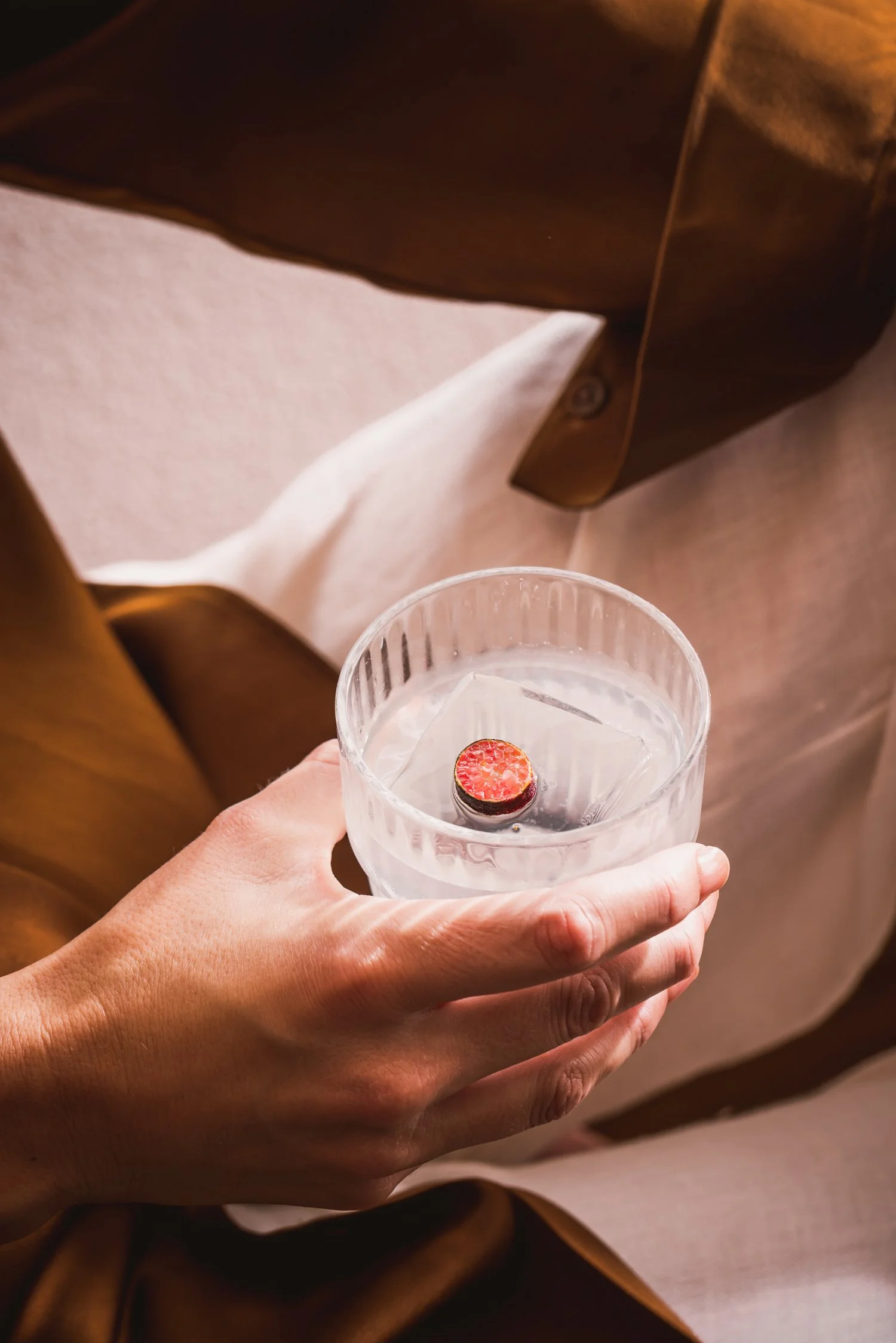 A person holding a glass of clear beverage with a sliced strawberry inside, viewed from above. The person is wearing a brown garment and a white shirt, and the background includes a brown fabric surface.
