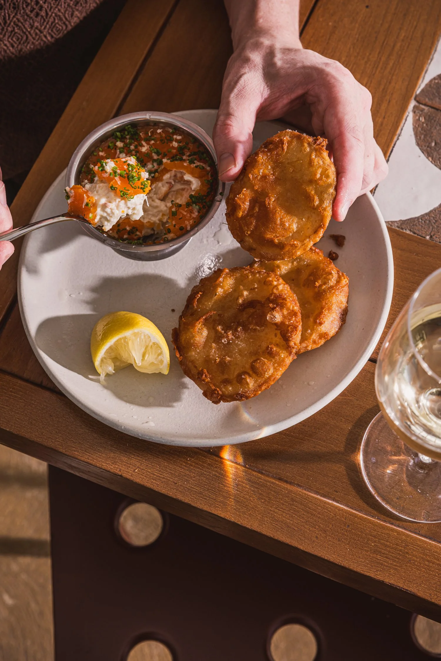 Fried fish fillets with lemon wedge and side of eggs, salmon, and cream in a small metal bowl, on a white plate with wooden table background.