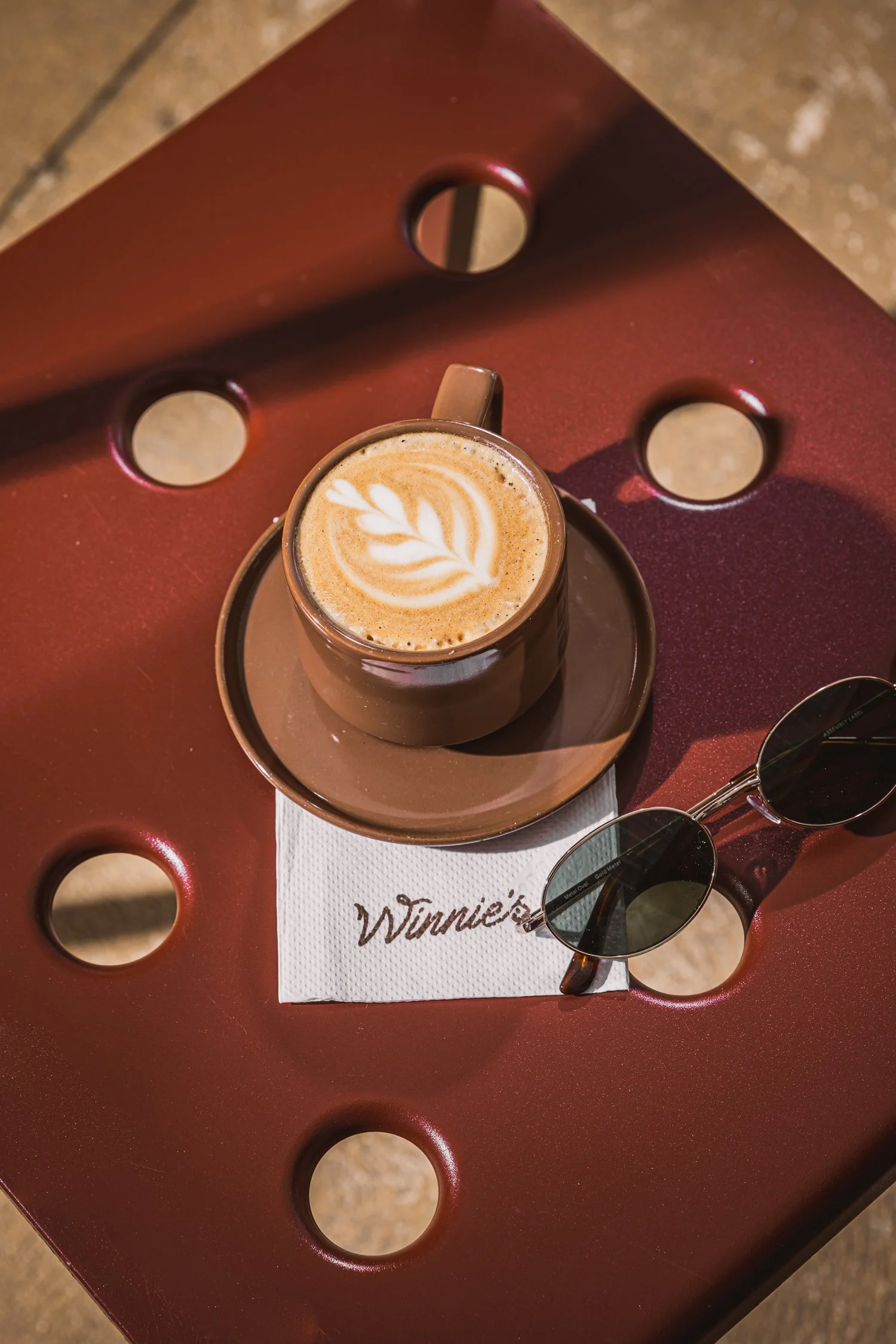 A cup of latte with latte art on top, placed on a saucer, with a pair of sunglasses and a white napkin with the word 'Winnie's' written on it on a red table with circular cutouts.