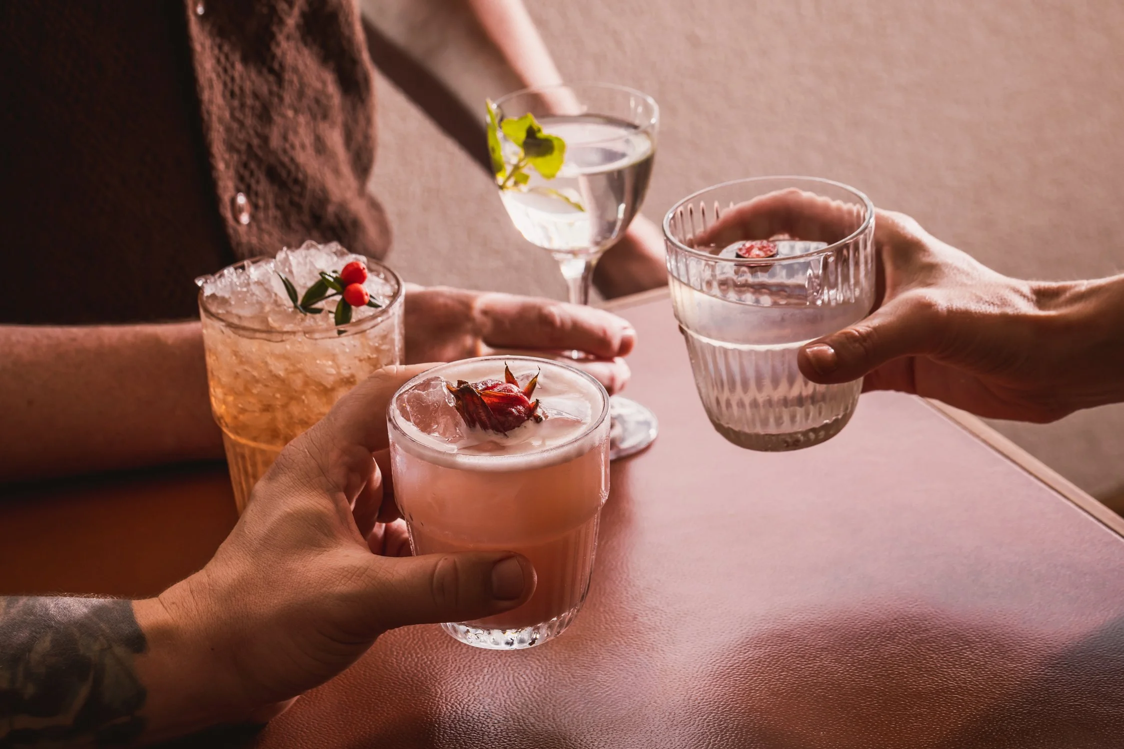 Four people are clinking glasses filled with pink cocktails, garnished with berries and herbs, in a toast celebration. The scene is warm and intimate, with a soft pink surface underneath.