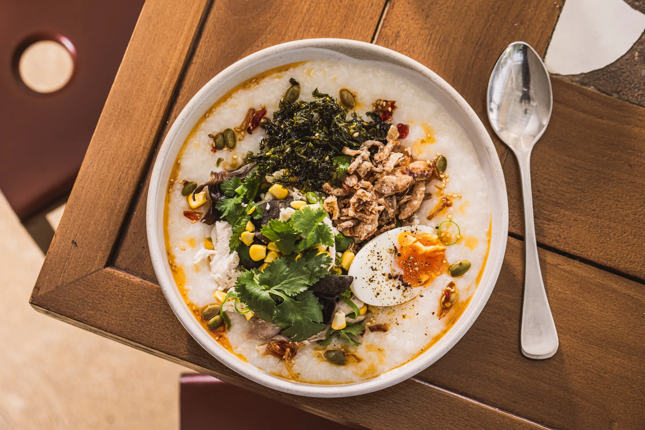 Bowl of Asian rice porridge topped with seaweed, chicken, corn, cilantro, pumpkin seeds, and a halved soft-boiled egg, placed on a wooden table with a spoon beside it.