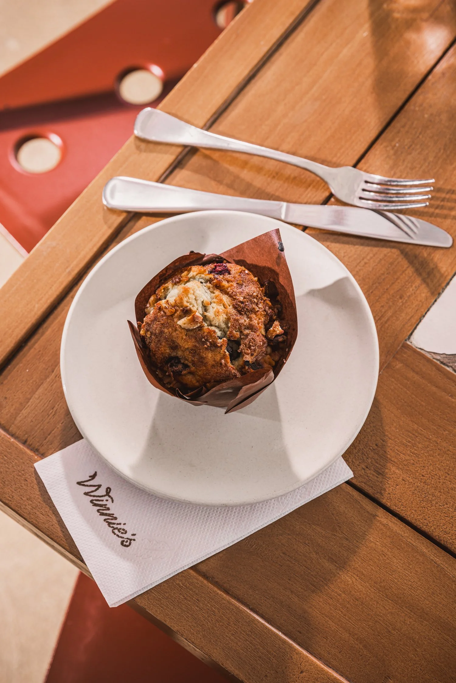 A muffin on a white plate with a napkin underneath, placed on a wooden table, with a fork and knife resting above the plate.