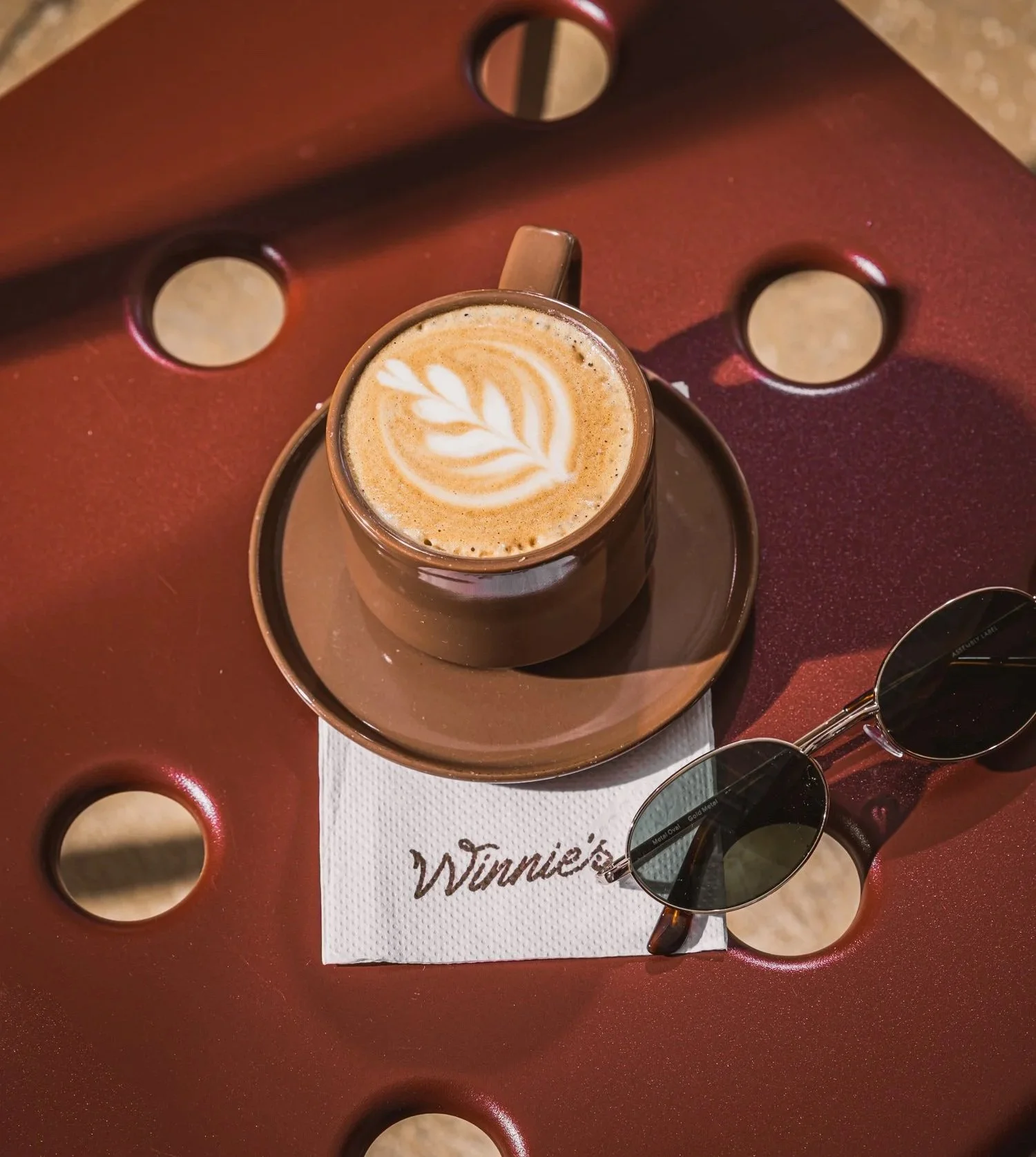 A cup of latte with foam art on a saucer, placed on a Wimmie's napkin on a brown, rounded table with circular cutouts, accompanied by sunglasses.