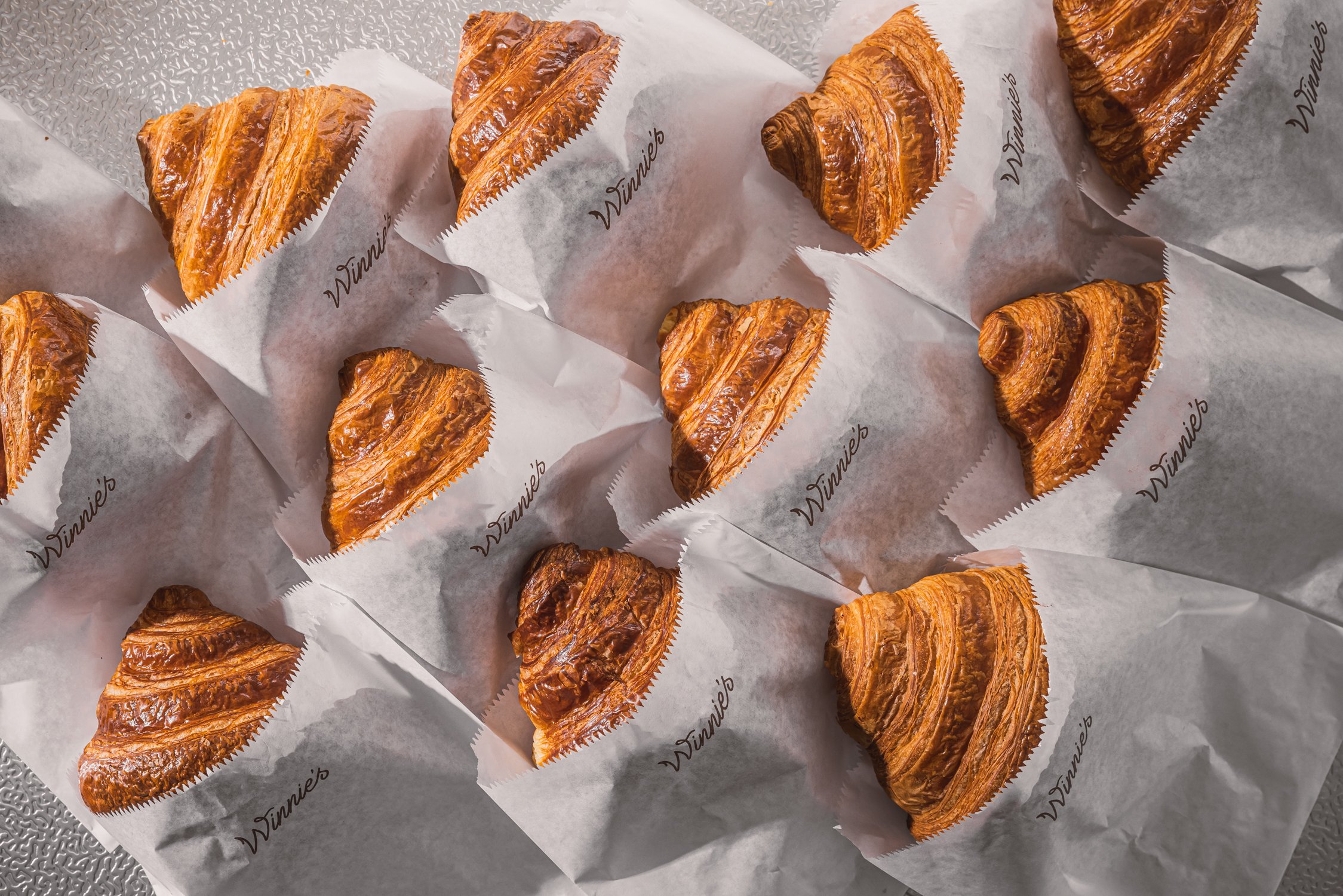 Several freshly baked croissants with flaky, golden-brown layers, individually wrapped in branded paper from 'Tinnie’s,' arranged on a textured surface.
