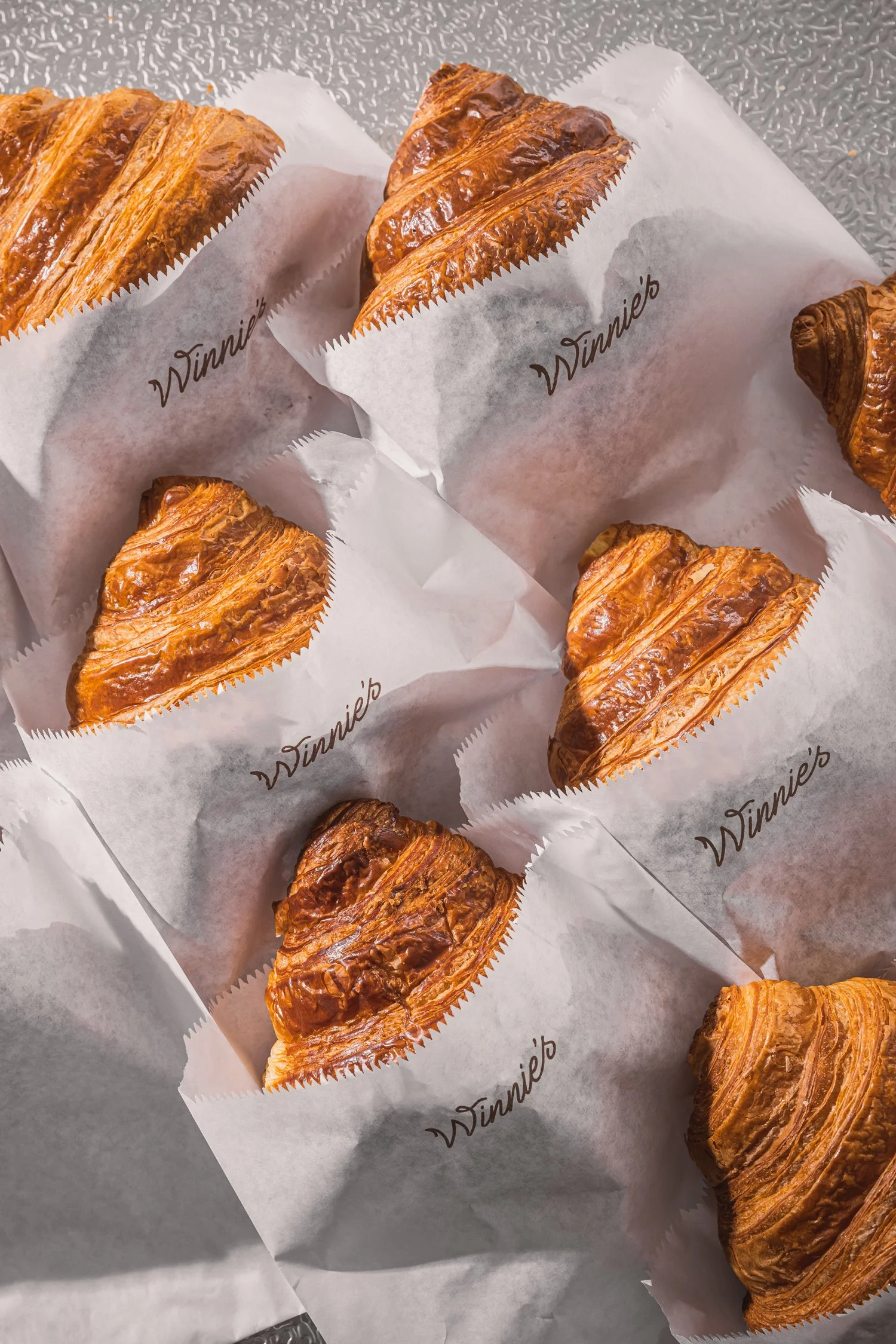 Several golden-brown croissants wrapped in white paper with the word 'Winnie's' printed on it, arranged on a textured gray surface.