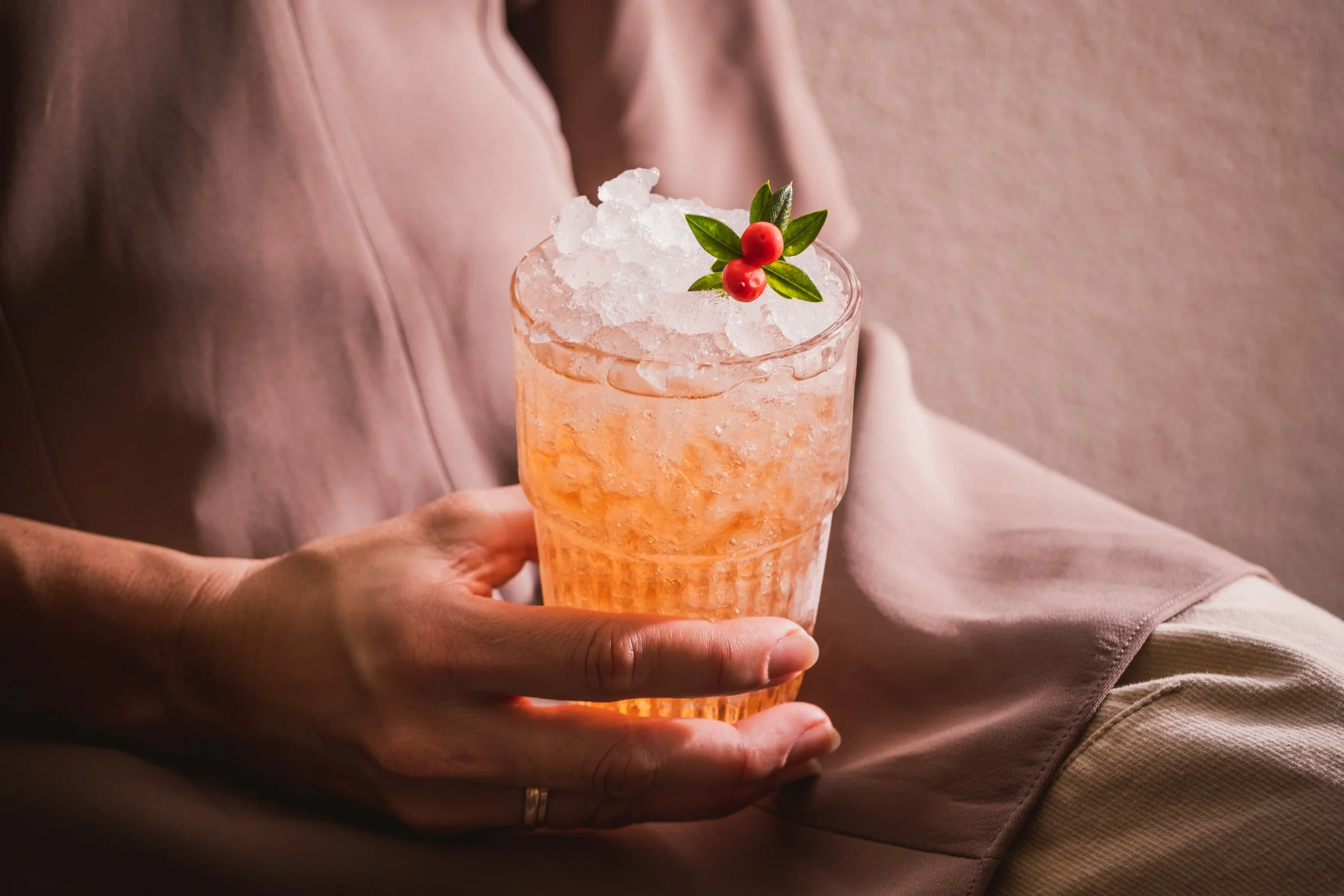 A person holding a glass of pink cocktail with ice, garnished with red berries and green leaves, wearing a light pink or beige long-sleeve shirt.