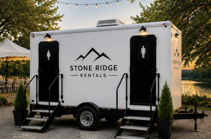 A white portable restroom trailer with Stone Ridge Rentals logo, black doors, stairs, and outdoor lighting, set up outdoors near trees and a body of water during sunset.