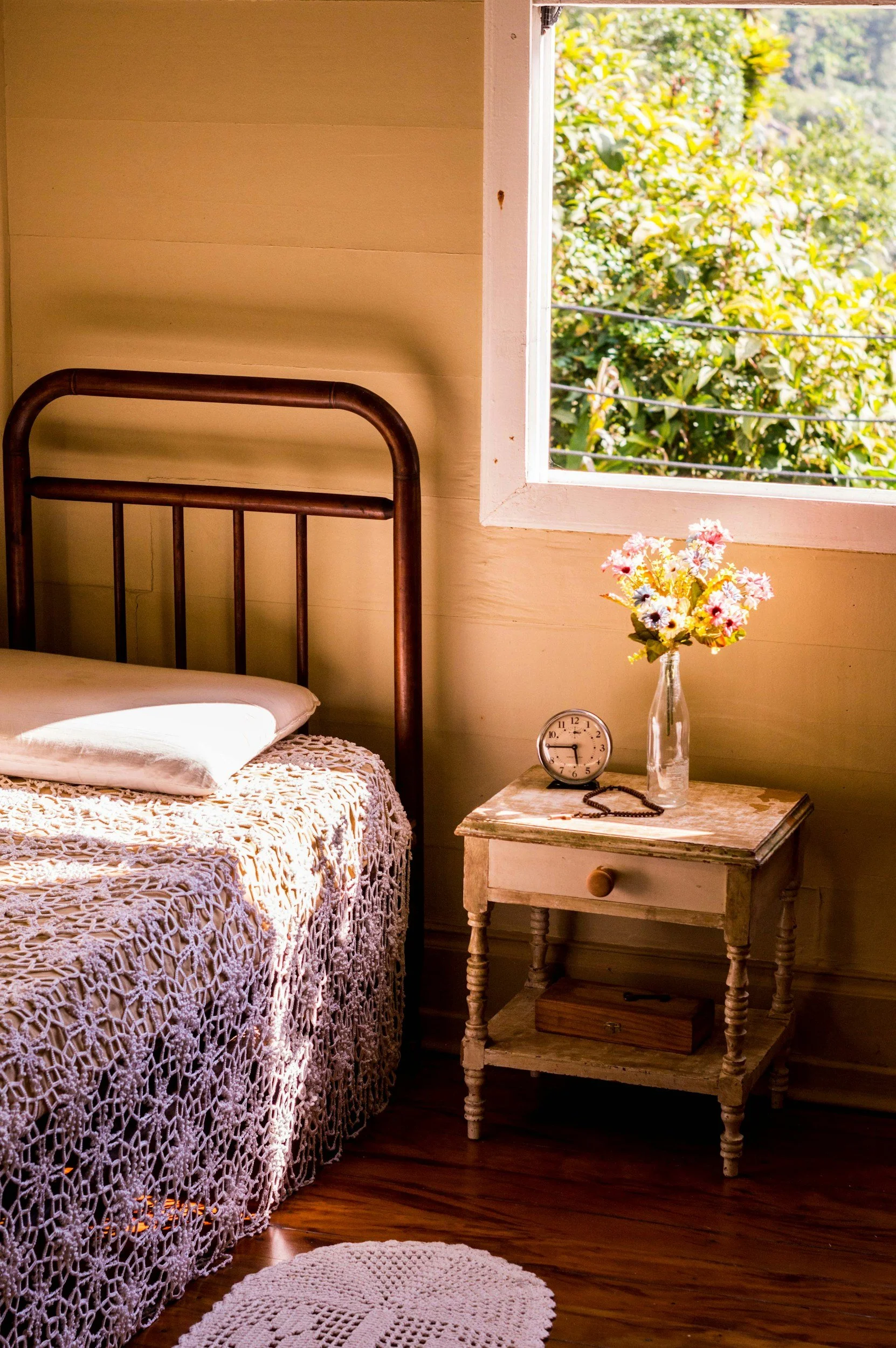 A cozy bedroom scene with a bed featuring a crocheted bedspread and a pillow, a vintage wooden nightstand holding a vase with a bouquet of flowers, a clock, and a rosary, and a window showing lush green foliage outside, with sunlight casting shadows inside.