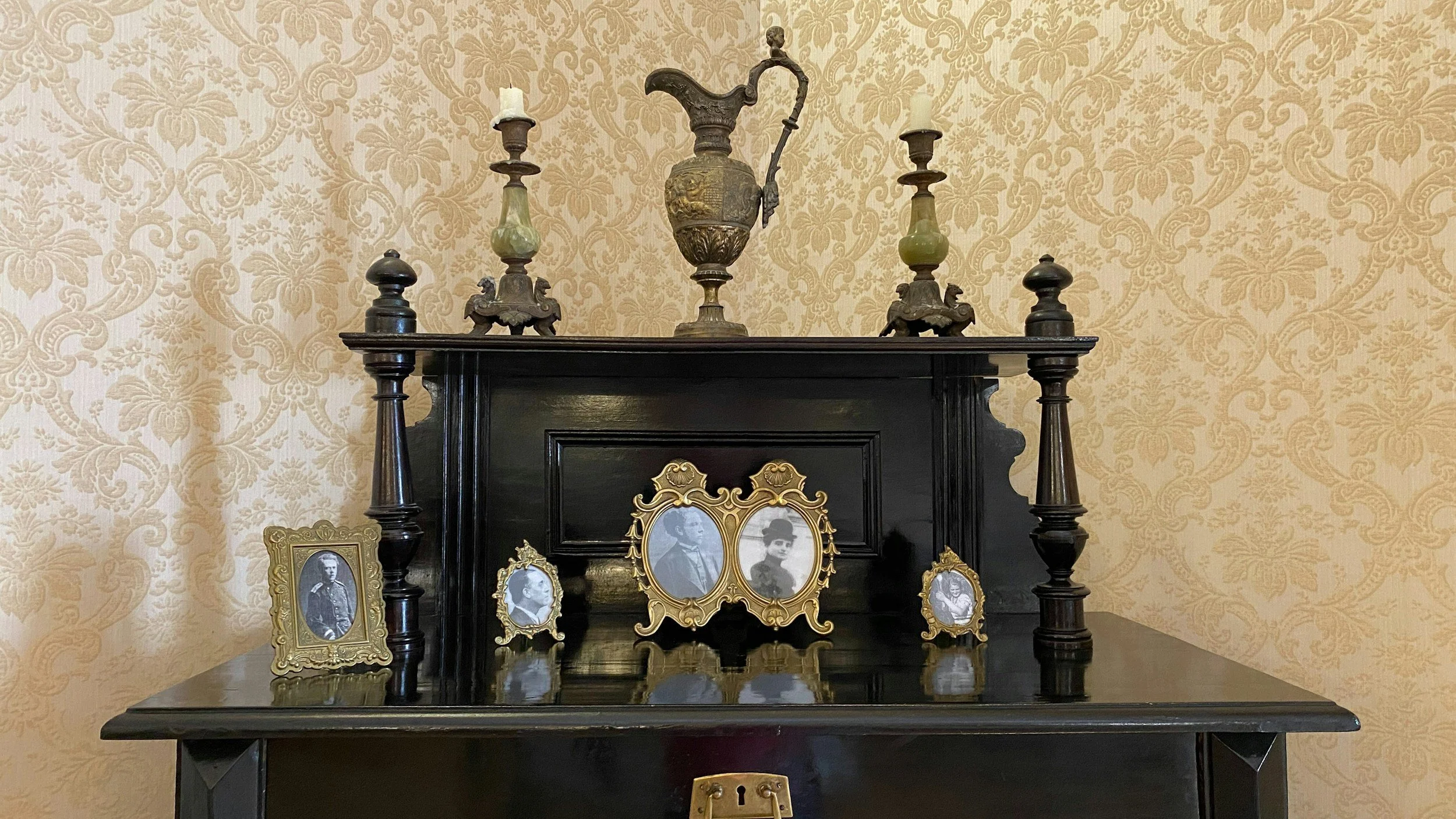 A black wooden cabinet with antique photo frames and a small ornate mirror on top, against a beige wallpapered wall with a floral pattern.