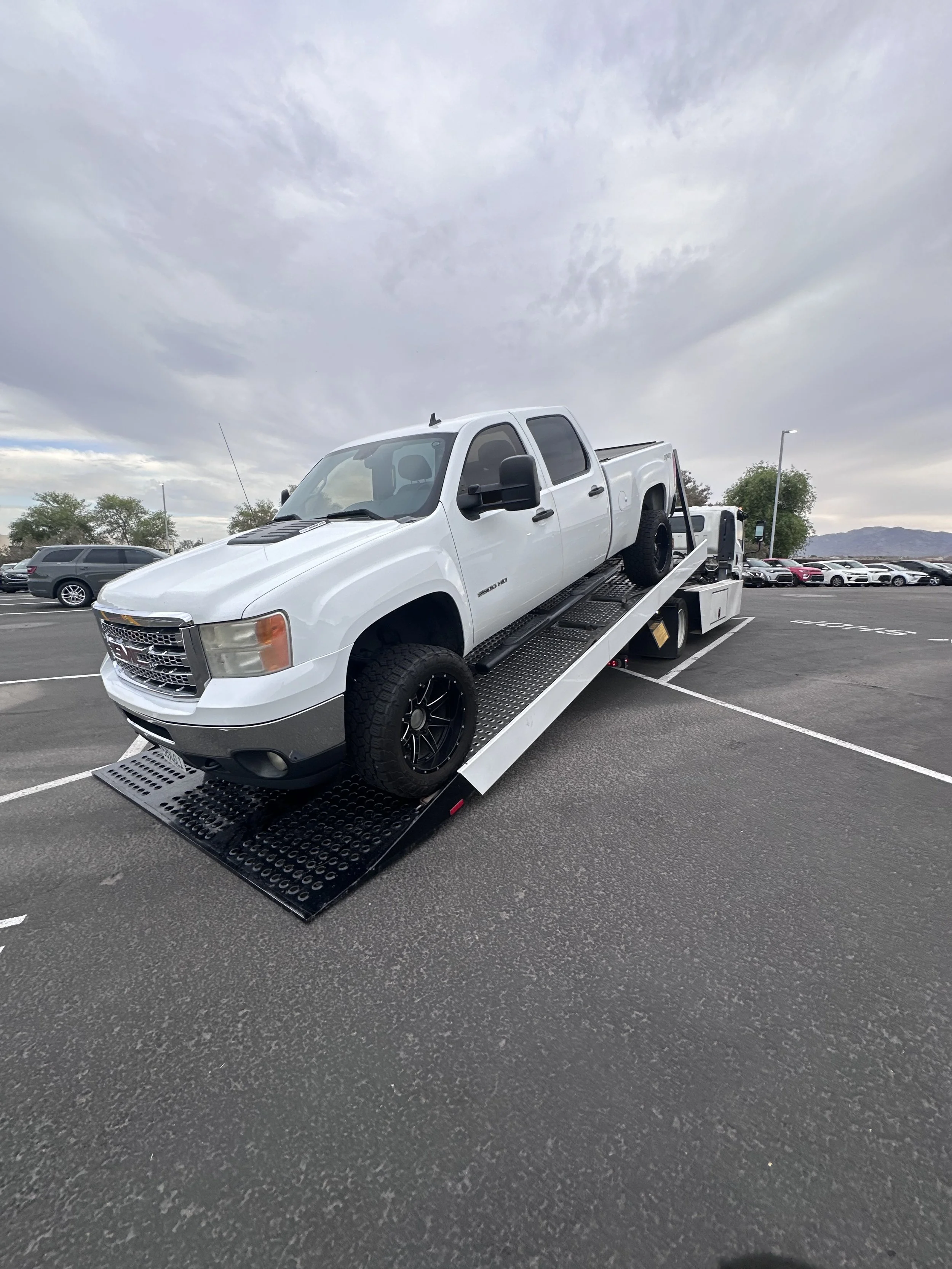 White pickup truck loaded onto a flatbed tow truck in a parking lot on a cloudy day.